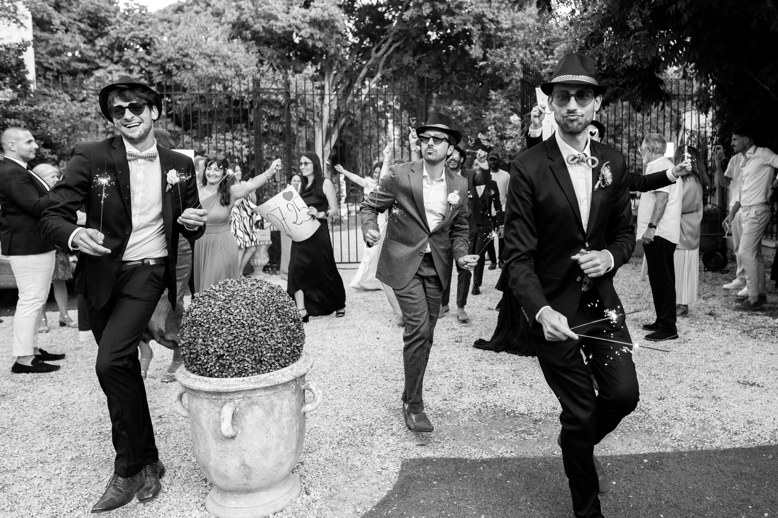 Groupe de personnes lors d'une fête ou célébration en plein air, certains dansant ou marchant, portant des costumes formels, avec des décorations et des confettis, dans un cadre naturel avec des arbres et une clôture.
