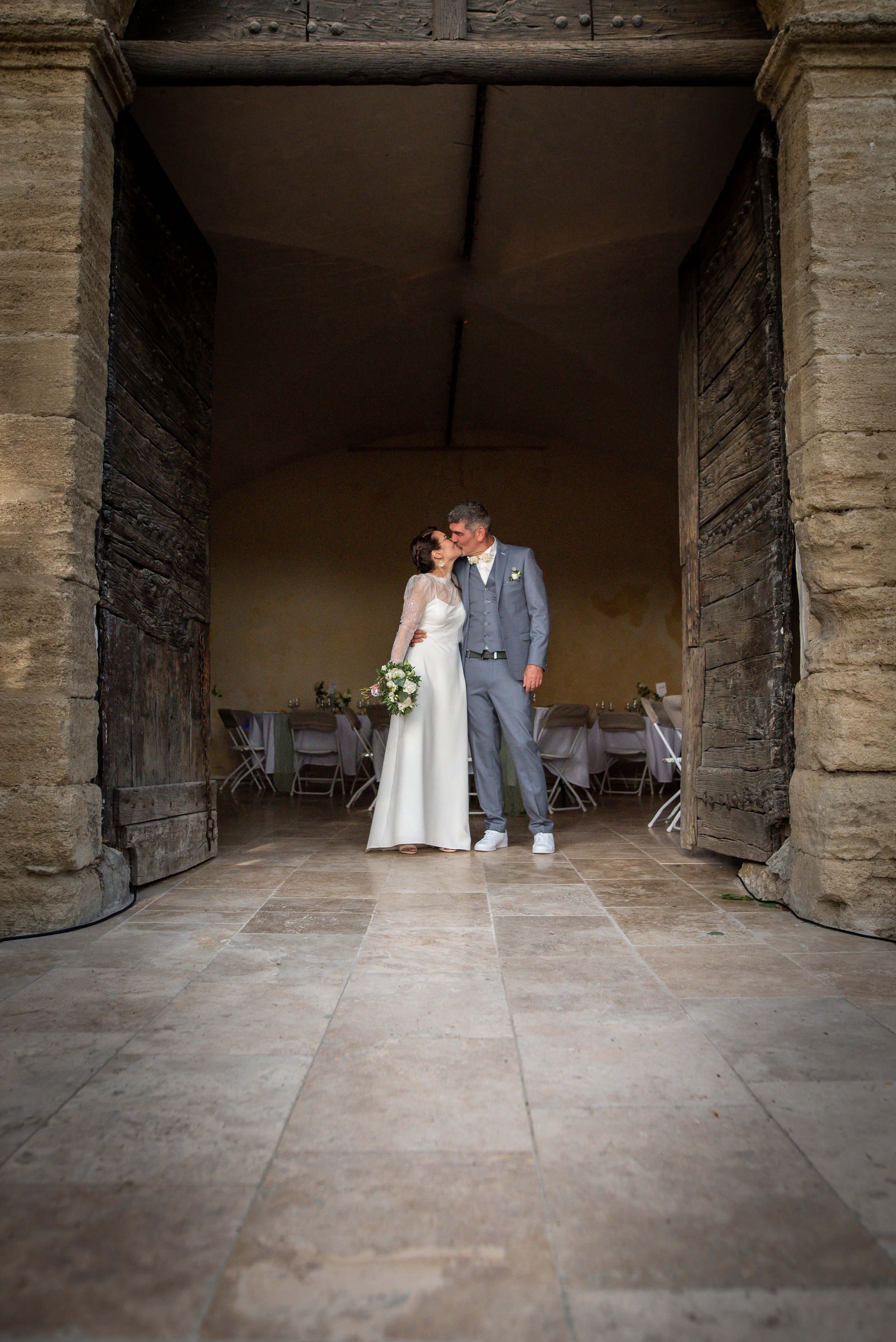 Un couple de mariés s'embrassant et s'embrassant sous une arche en pierre dans une salle de mariage avec des tables et des chaises en arrière-plan.