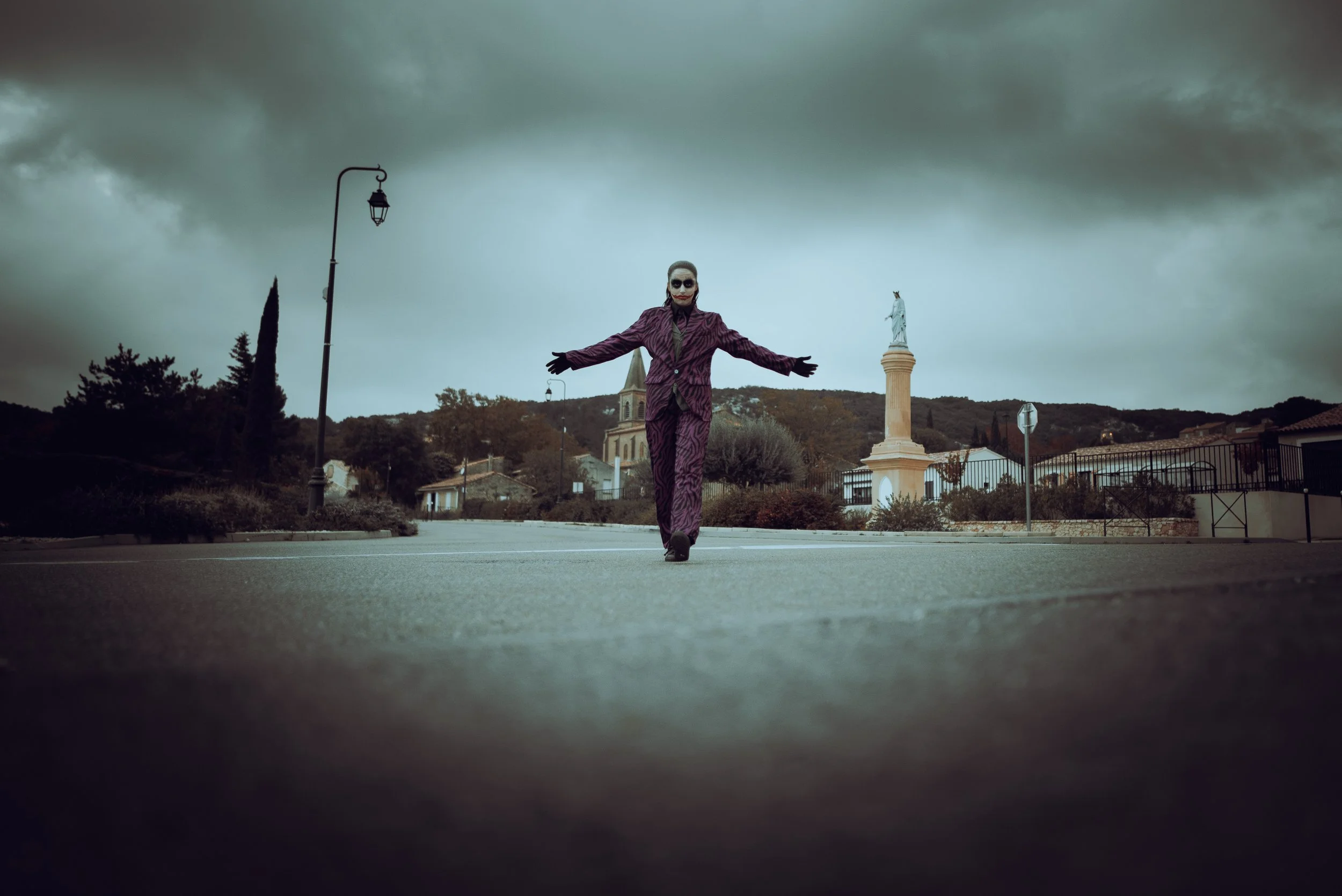 Une personne déguisée en le Joker, avec un costume violet à motifs, un visage maquillé en clown, marchant au centre d'une rue vide, sous un ciel nuageux, avec une statue de la liberté en arrière-plan. photographe mariage Nimes delor alexandre