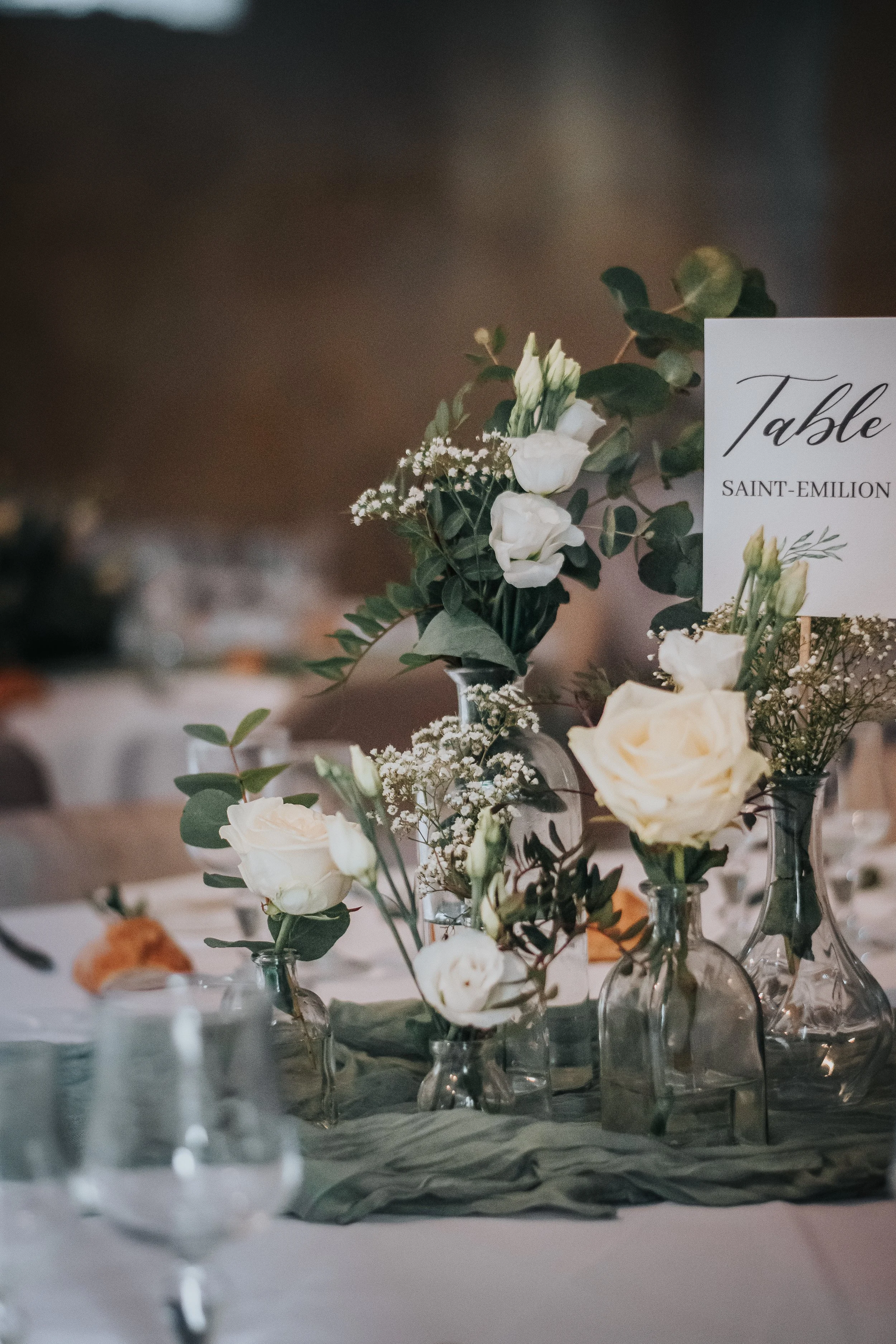 Central de fleurs blanches avec vases en verre, table de mariage avec une carte indiquant 'Table Saint-Emilion'.