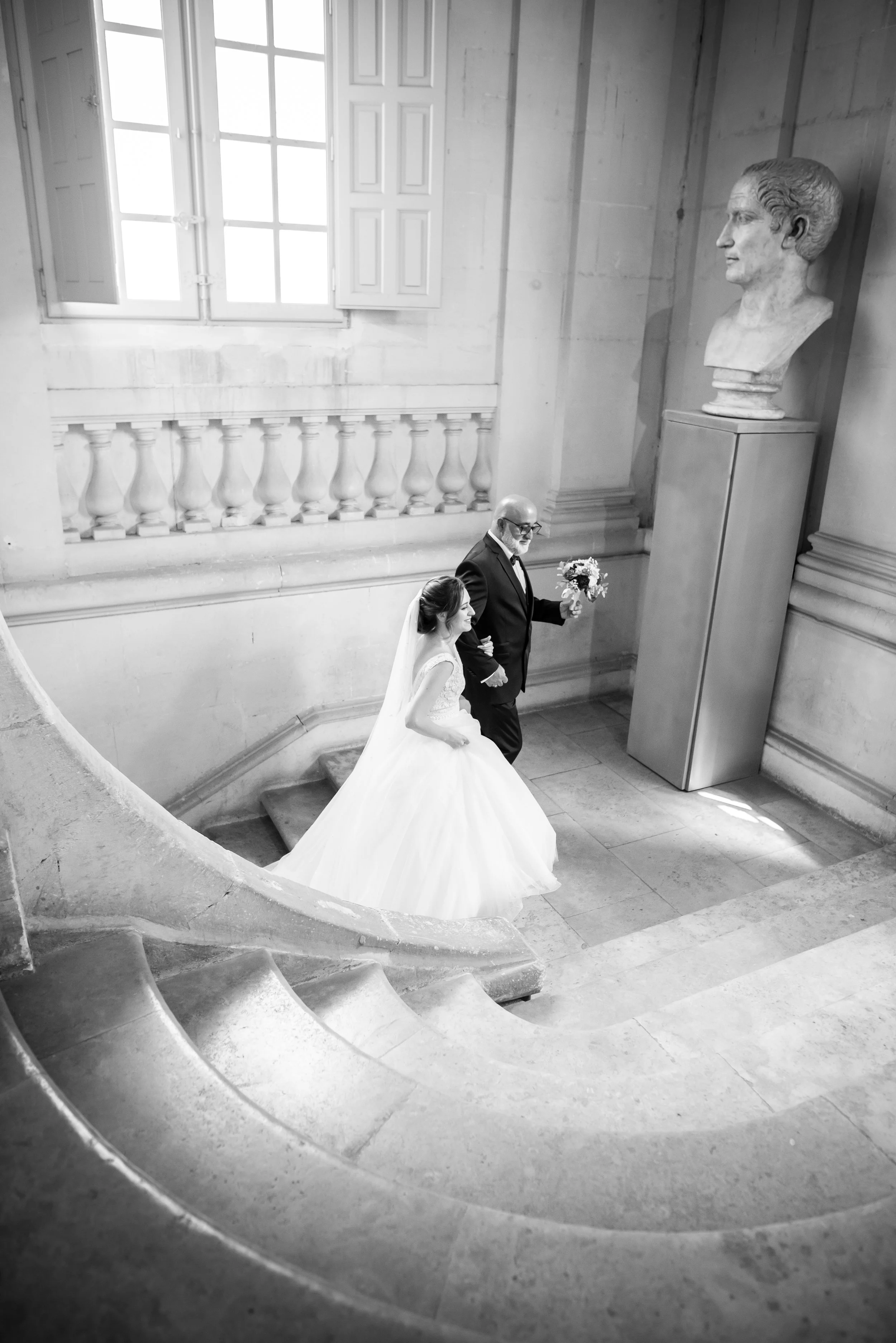 Une mariée en robe blanche et un homme en costume noir, probablement son père, descendent un escalier en pierre dans un bâtiment historique, avec une grande statue de profil sur un piédestal et une fenêtre laissant passer la lumière.