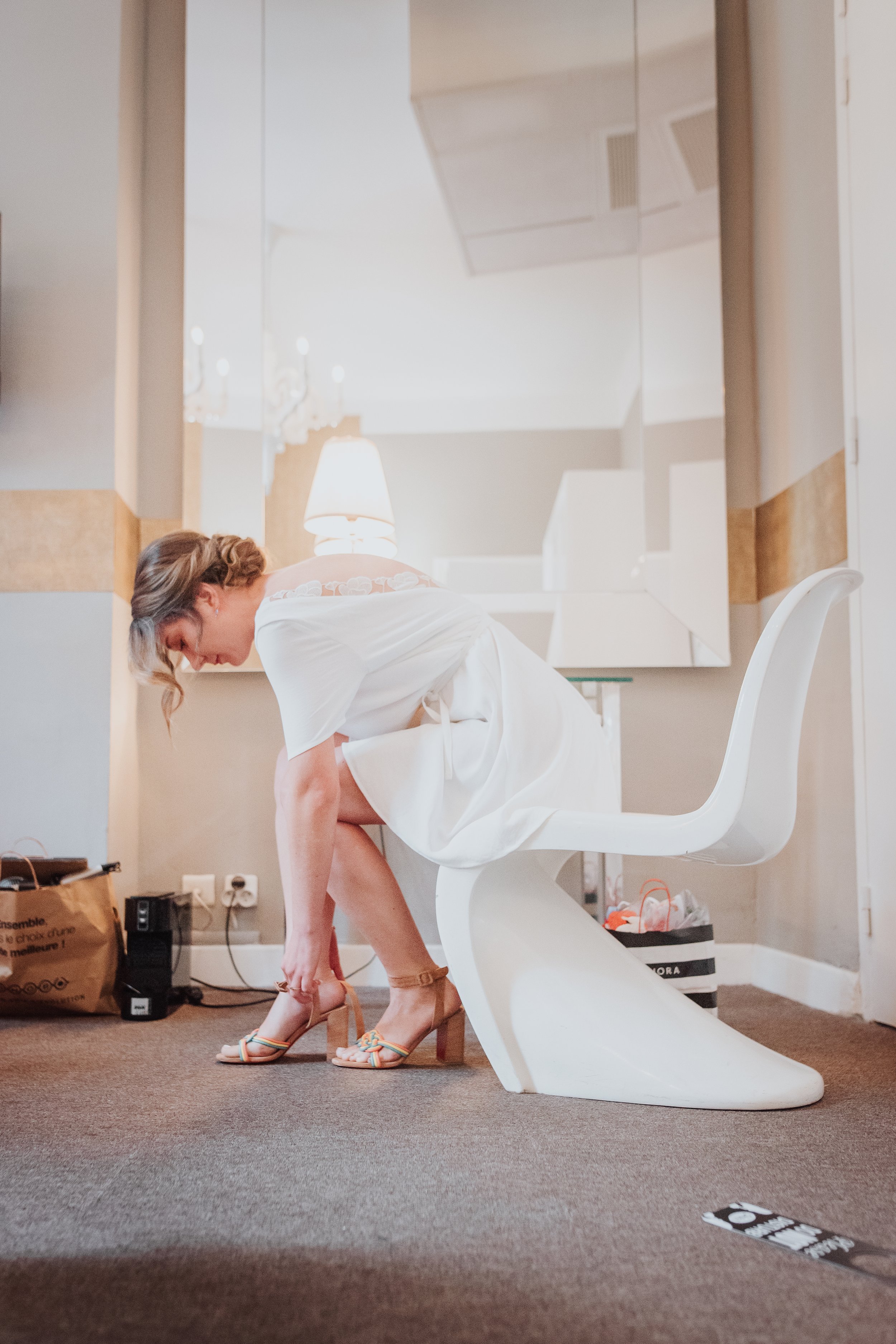 Une femme en robe blanche essayant de mettre des sandales à talons hauts dans une pièce lumineuse, près d'un miroir mural.