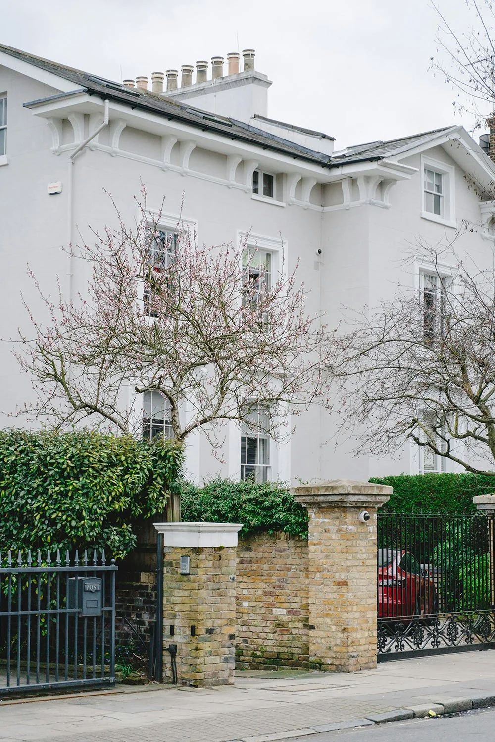 A white multi-story house with small windows, a gated entrance, and a brick wall. Two leafless trees are in front of the house, and a red car is parked behind the gate.