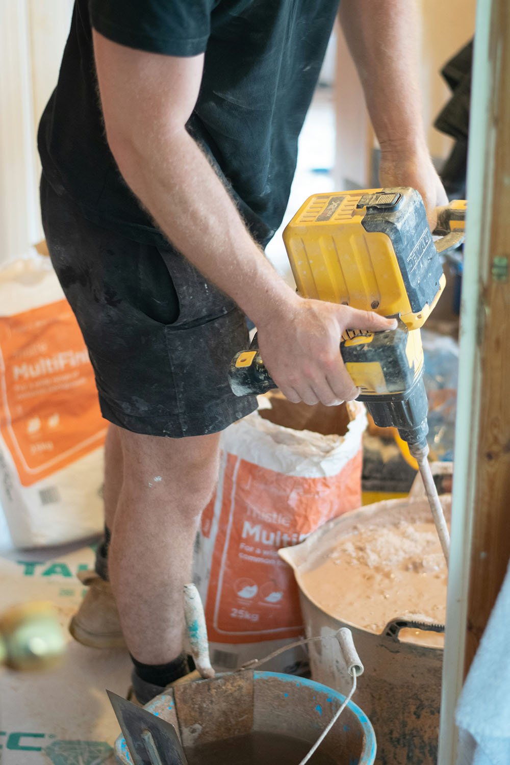 A person using a yellow power drill while doing construction work in a room with various construction materials and bags of mortar or drywall compound.