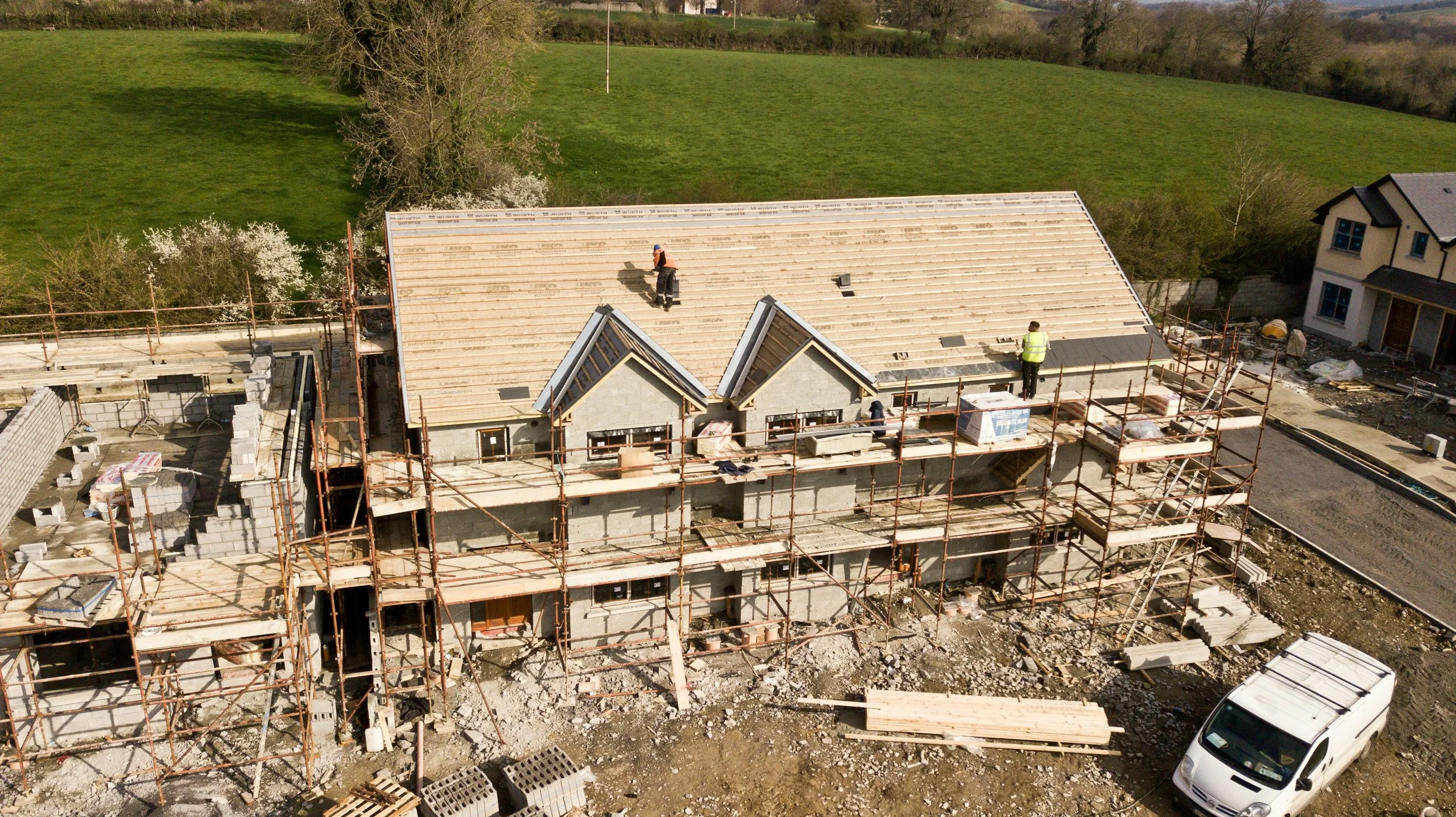 Construction site with workers building a multi-story residential or commercial building, scaffolding surrounding the structure, and a green field with trees in the background.