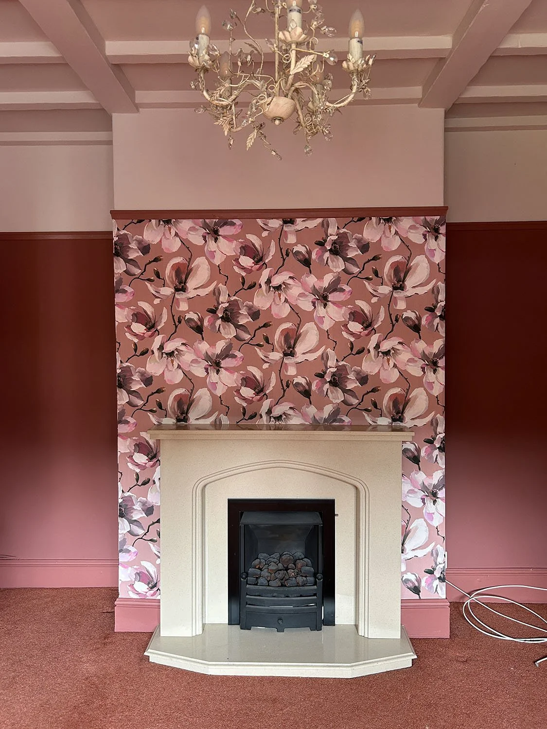 A room with a pink floral wallpaper above a fireplace with a cream mantel and a black fire insert, topped with a chandelier and pink walls on either side.