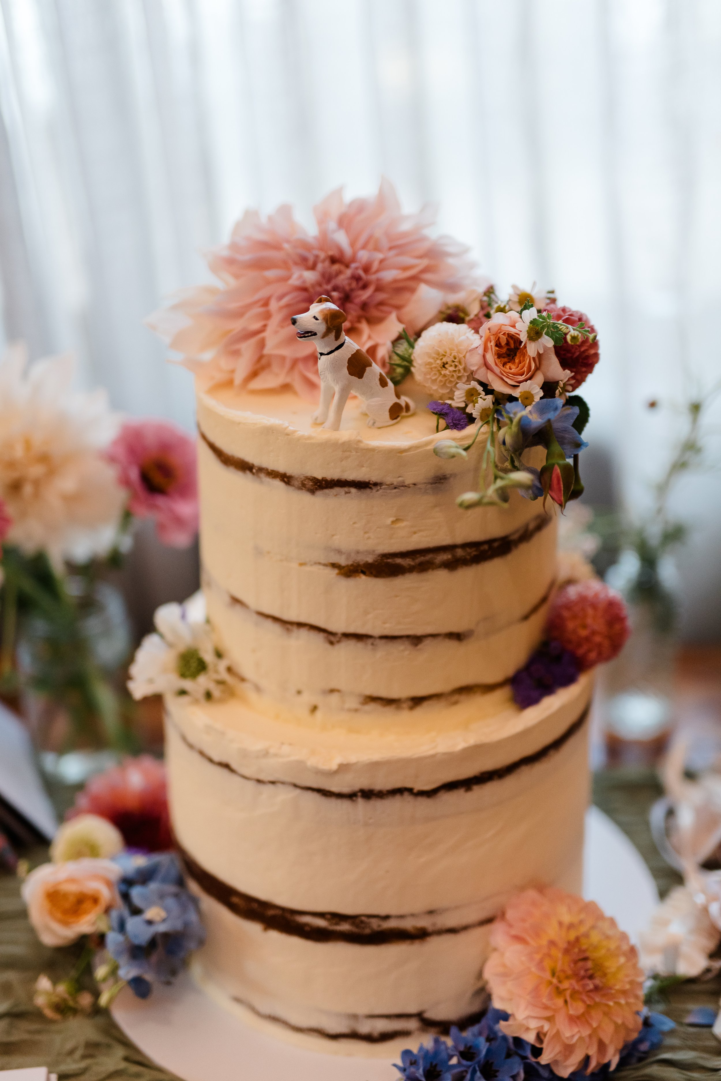 A wedding cake with two tiers decorated with flowers and a dog figurine on top, featuring a semi-naked frosting style and surrounded by additional flowers.