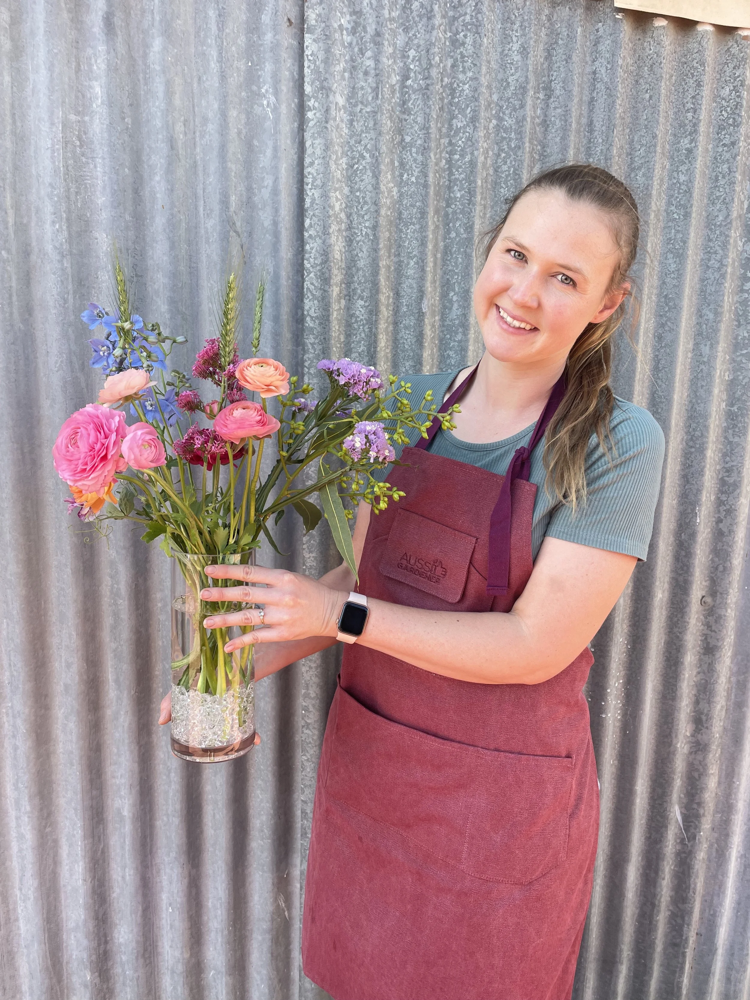 Smiling woman wearing a teal shirt and maroon apron holding a colorful bouquet of flowers in a clear glass vase against a corrugated metal background.