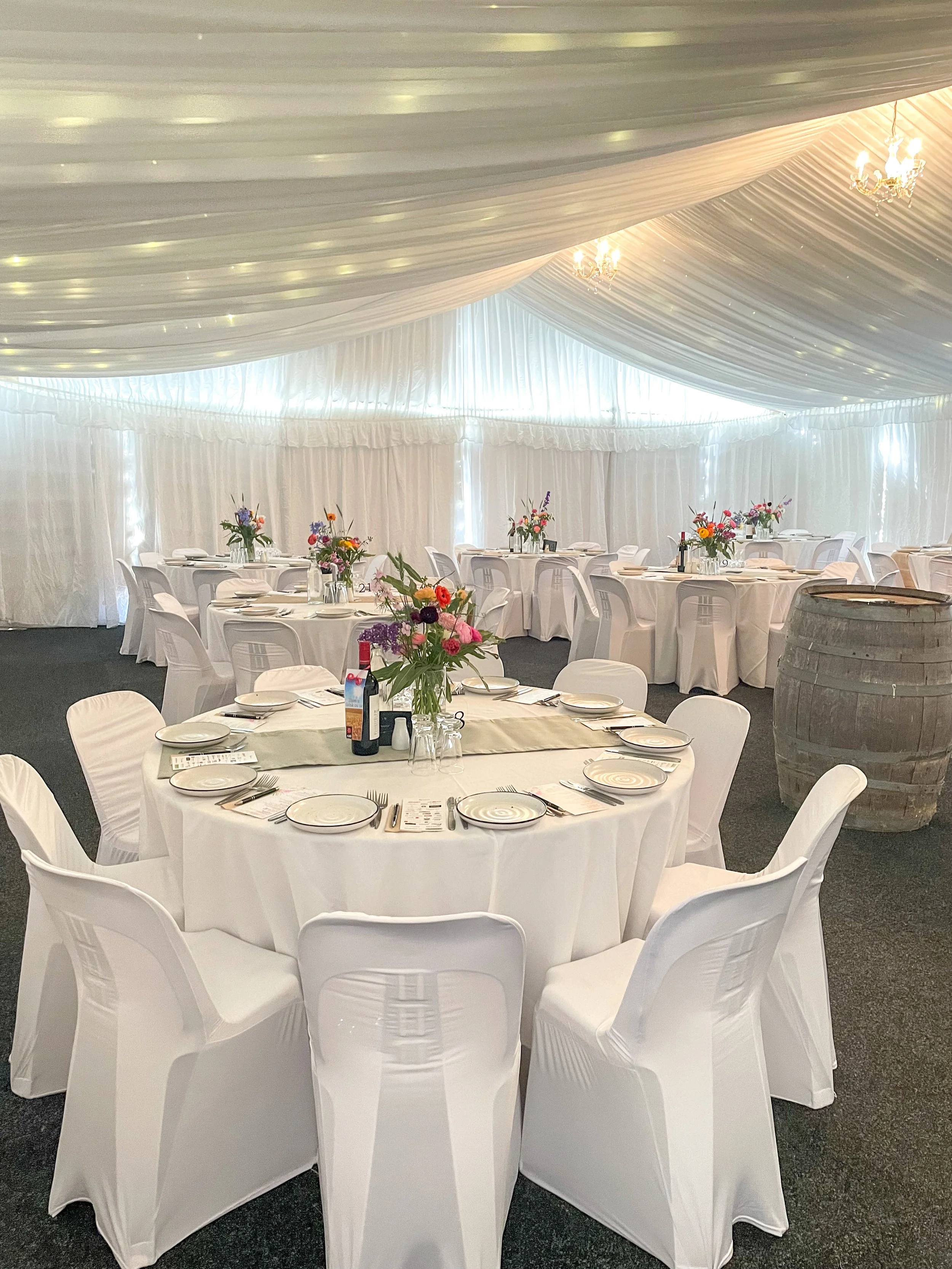 Elegant banquet hall set up with round tables covered with white tablecloths, pink and purple floral centerpieces, white chairs, and a decorated ceiling with hanging chandeliers.