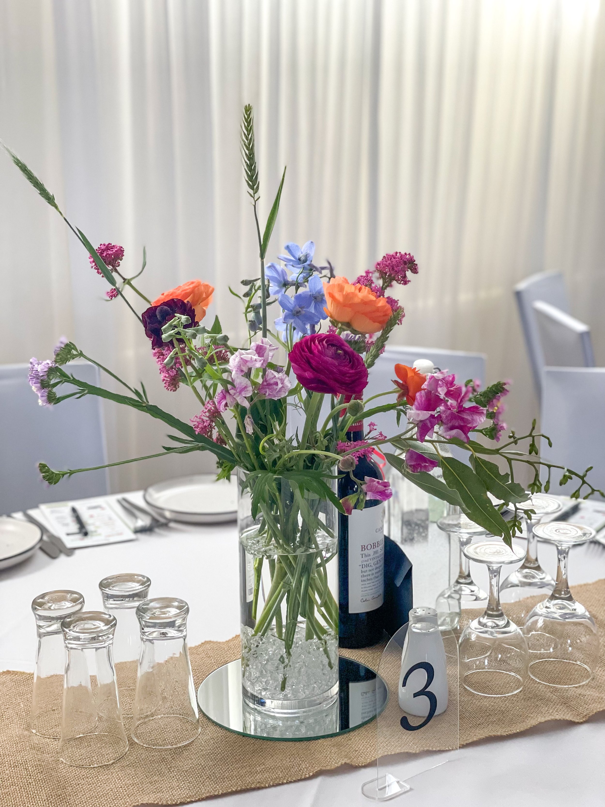 A table centerpiece with a colorful bouquet of mixed flowers in a glass vase on a mirror with a burlap table runner, surrounded by upside-down wine glasses and a small table number.