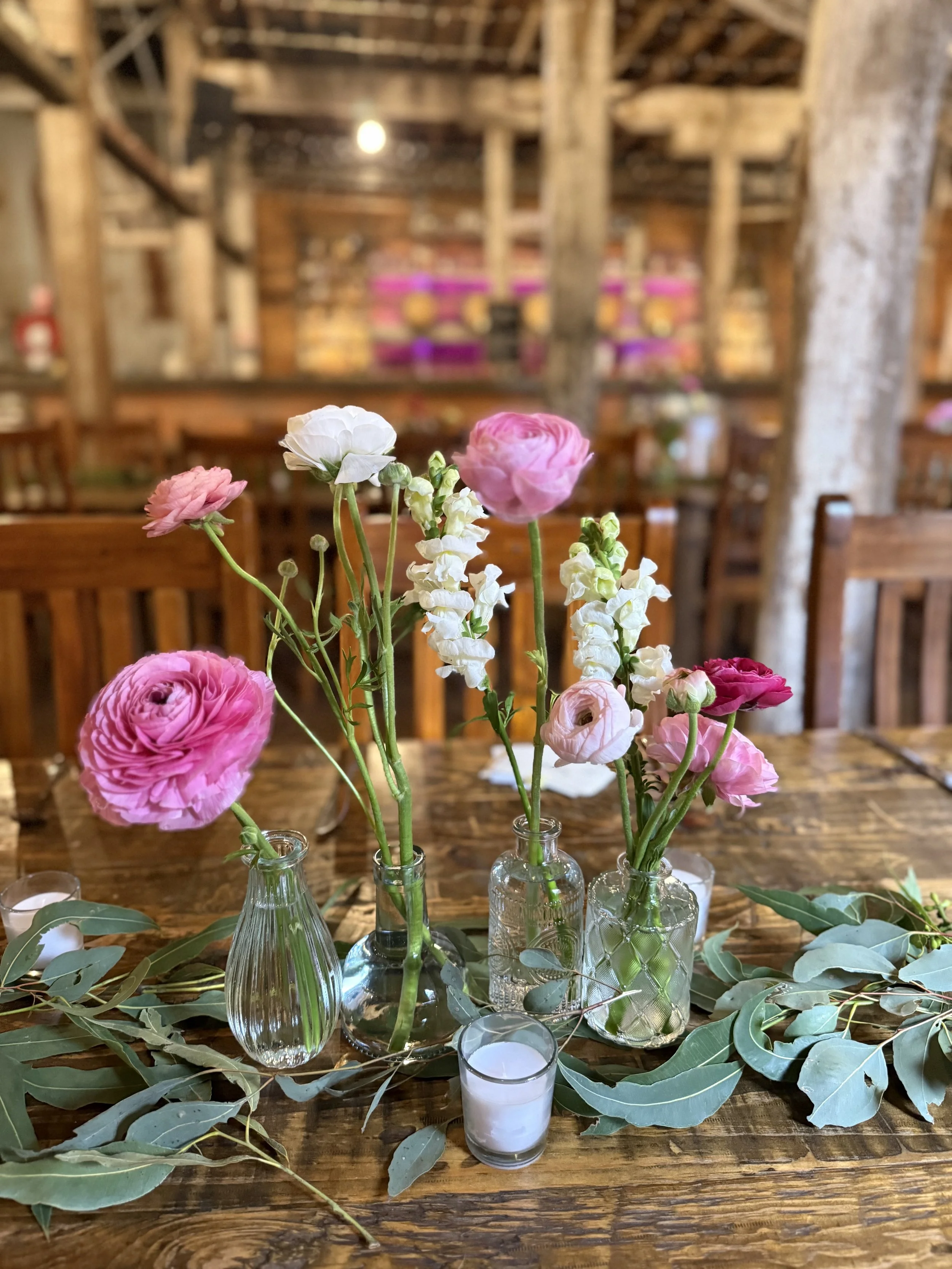 A rustic wooden table with small glass vases holding pink, white, and purple ranunculus flowers, surrounded by eucalyptus leaves and a small white candle, inside a warmly lit barn-like venue.