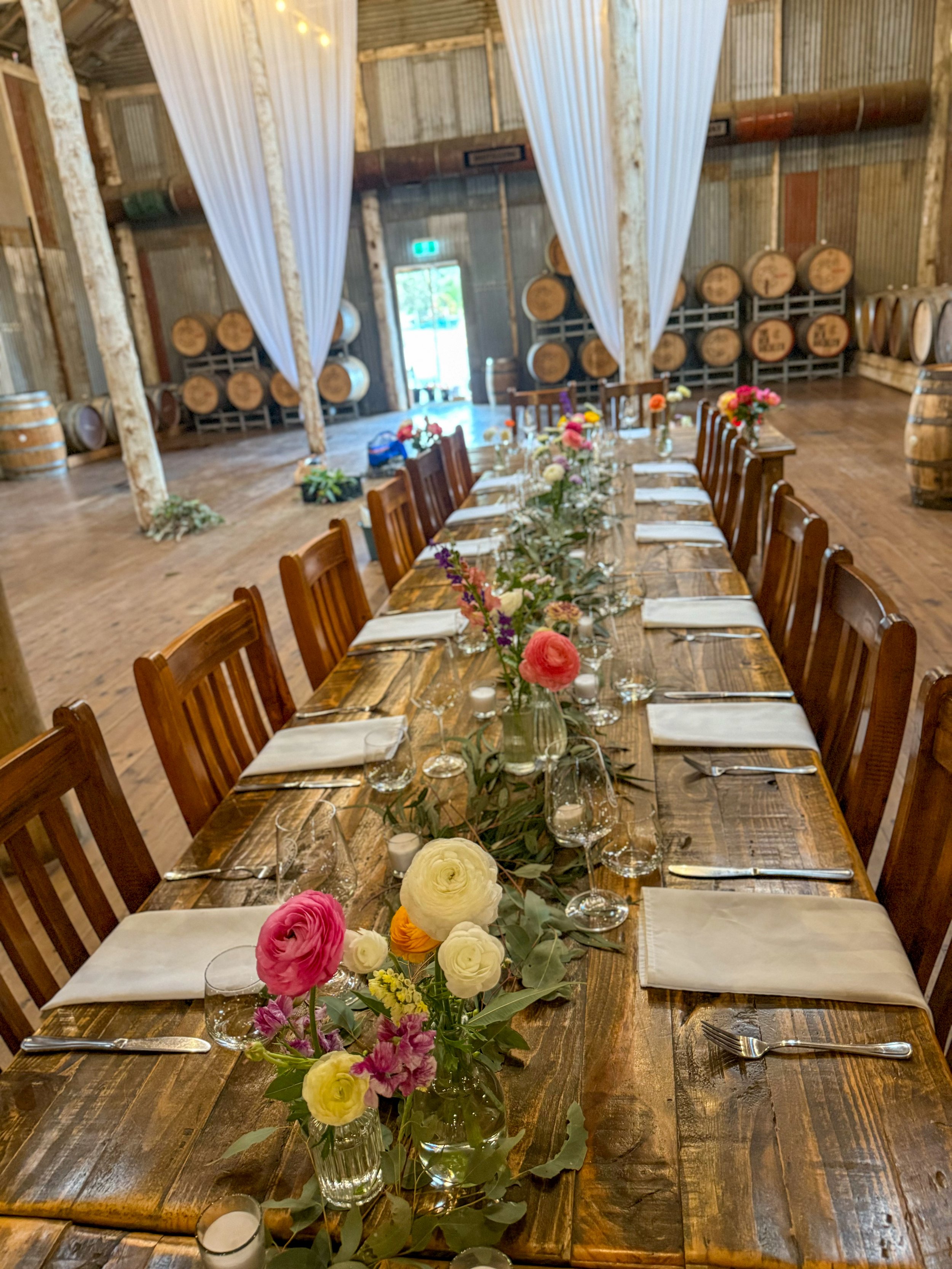 Long wooden banquet table set for a formal event with floral centerpieces, white napkins, glasses, and silverware in a rustic winery with barrels in the background.