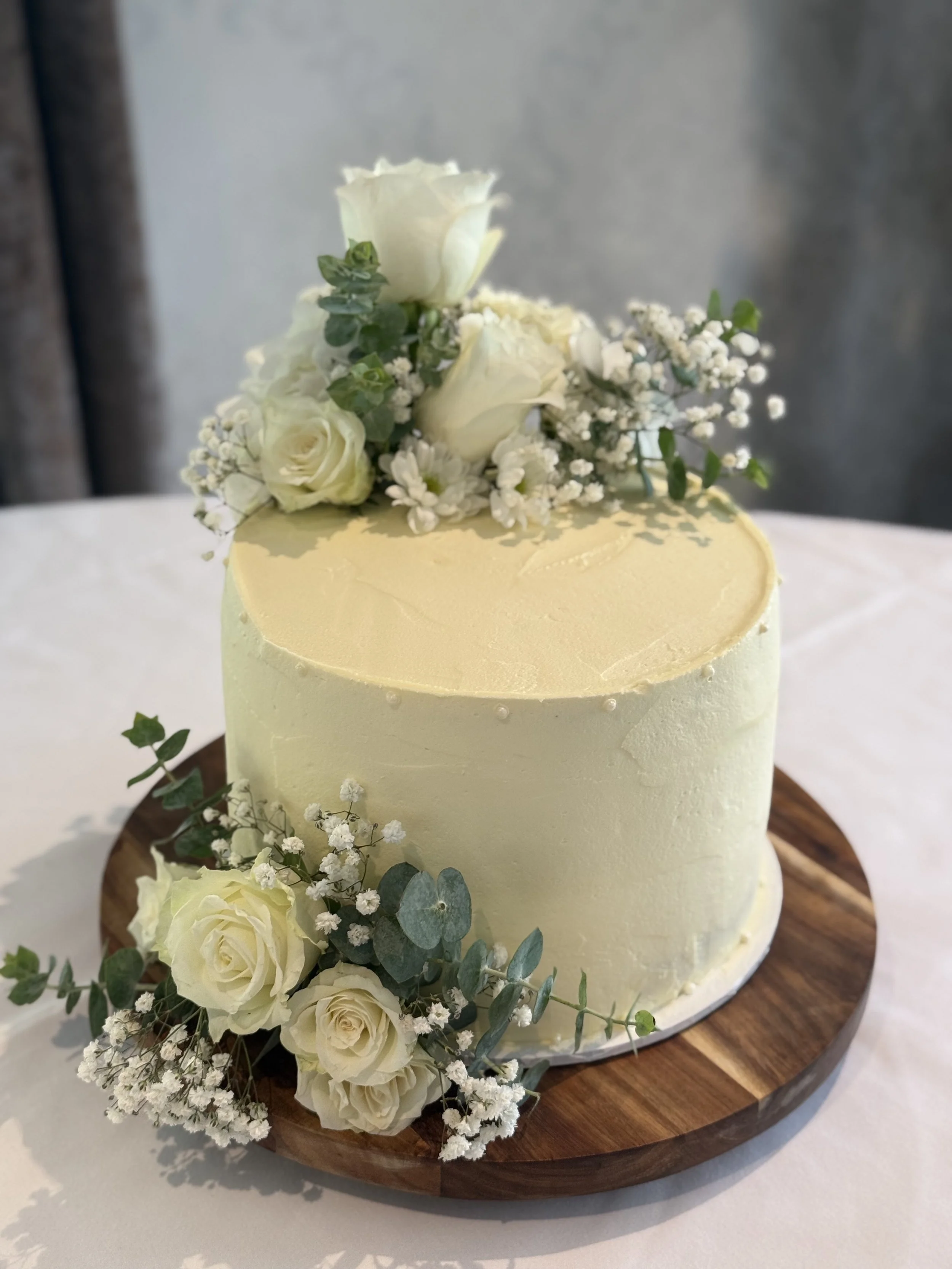 Cream-colored wedding cake decorated with white roses and baby's breath, placed on a wooden cake stand.