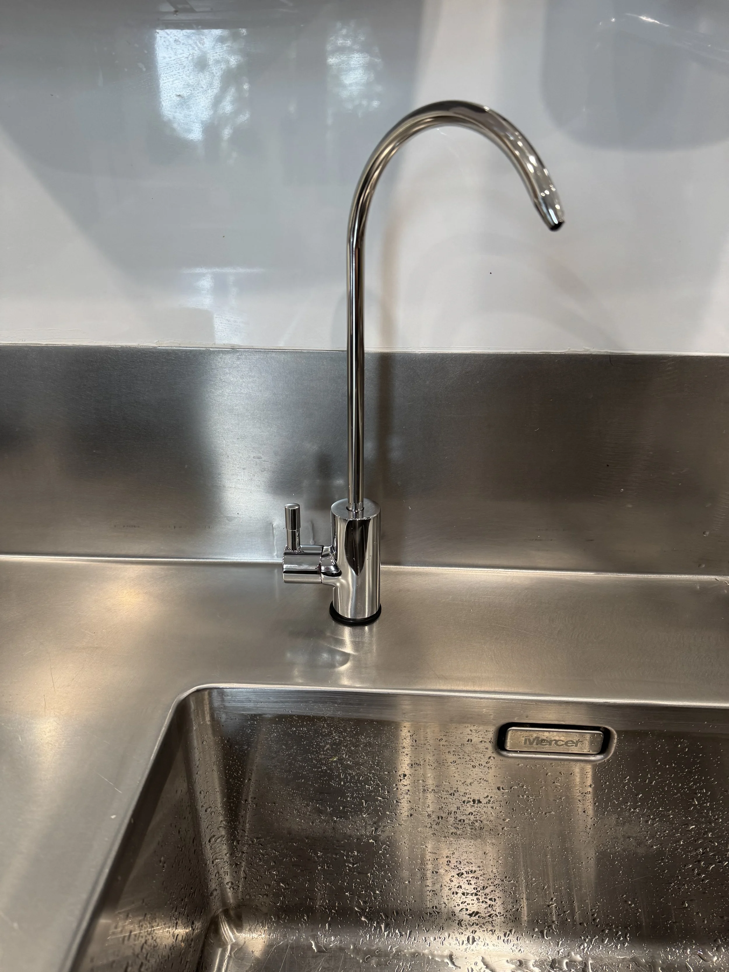Stainless steel kitchen sink with a tall, curved faucet and a soap dispenser, with water droplets inside the sink.