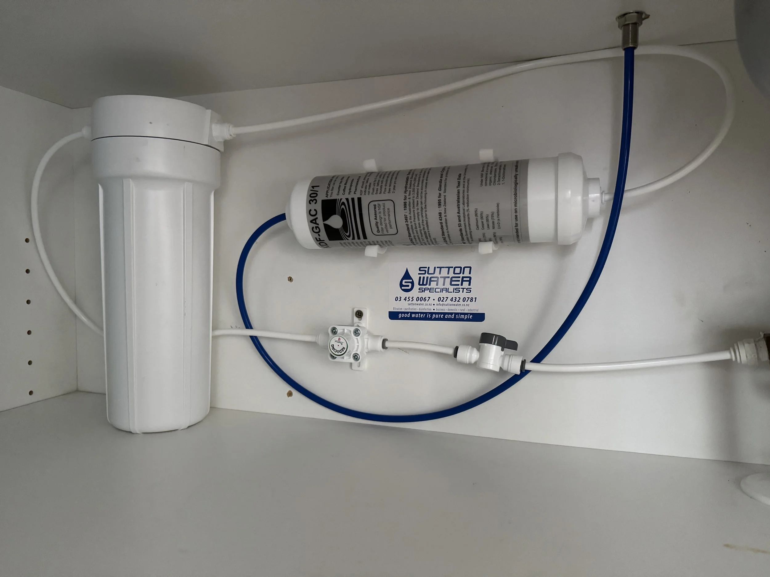 Under-sink water filtration system with a white filter housing, a reverse osmosis membrane, blue and white tubing, and a pressure gauge.