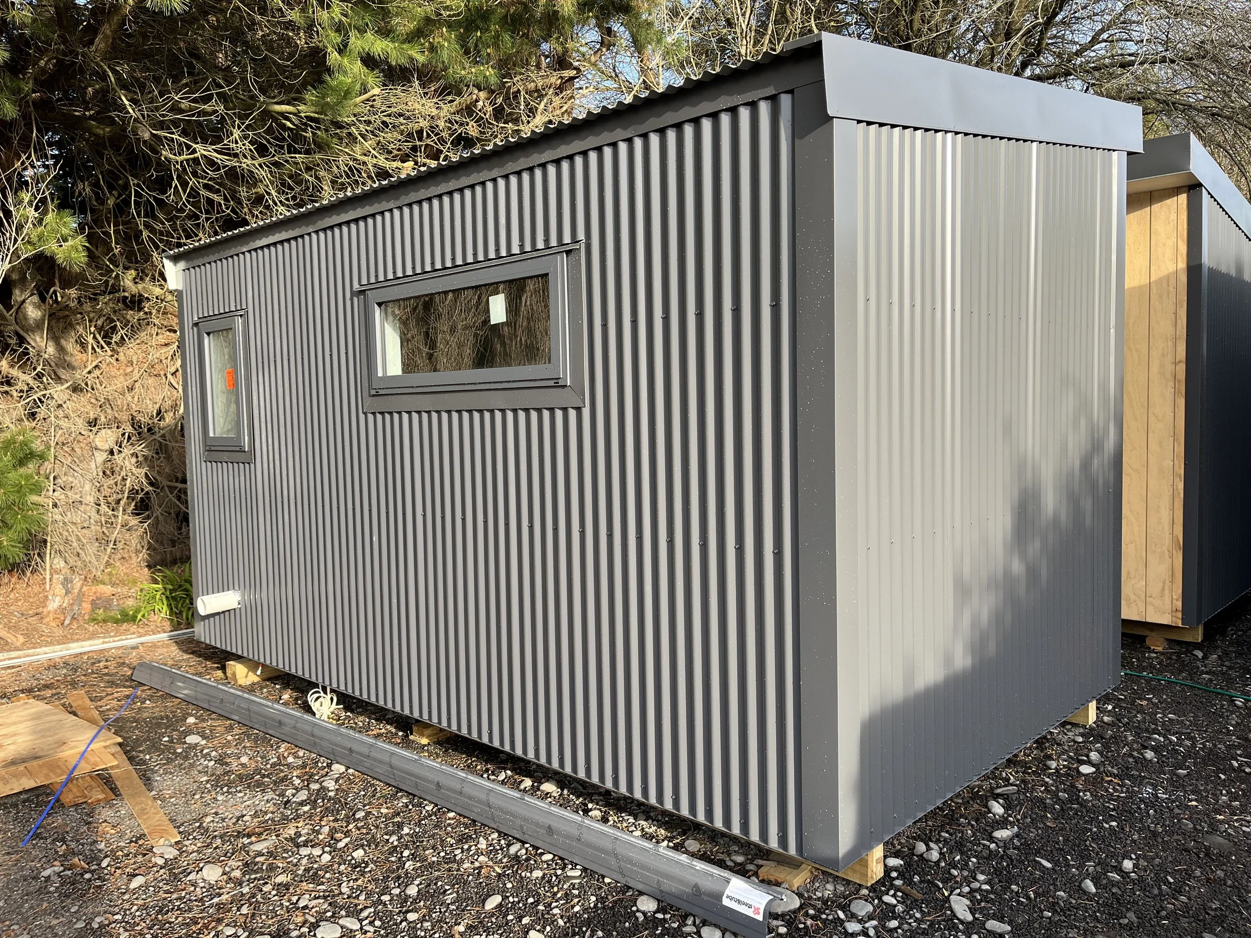 Small modern building with gray corrugated metal siding, windows, and a wooden section, situated on uneven ground with construction materials around, near trees.
