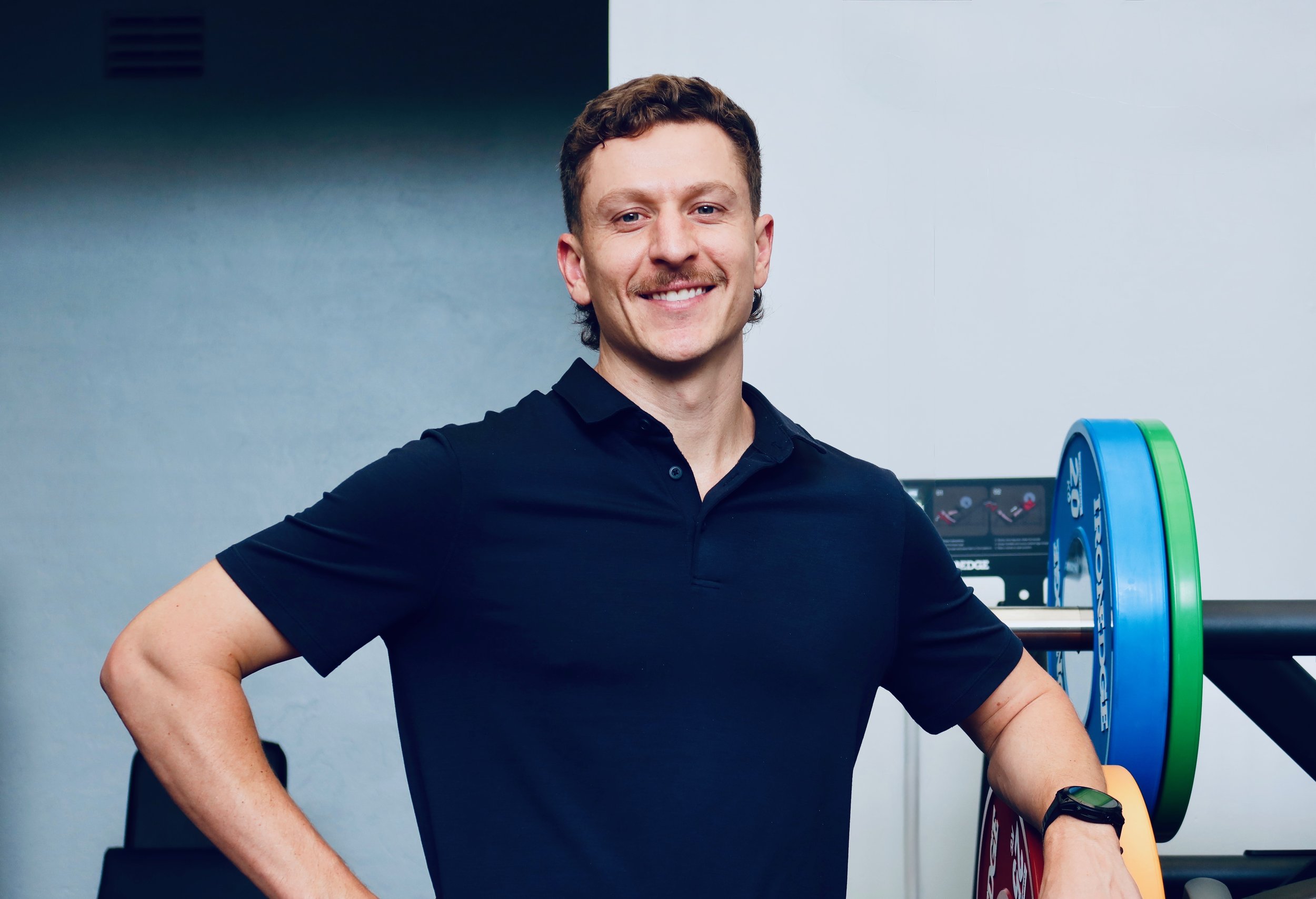 A man with brown hair and a mustache smiling, wearing a navy blue polo shirt, standing in a gym with colorful weight plates and workout equipment in the background.