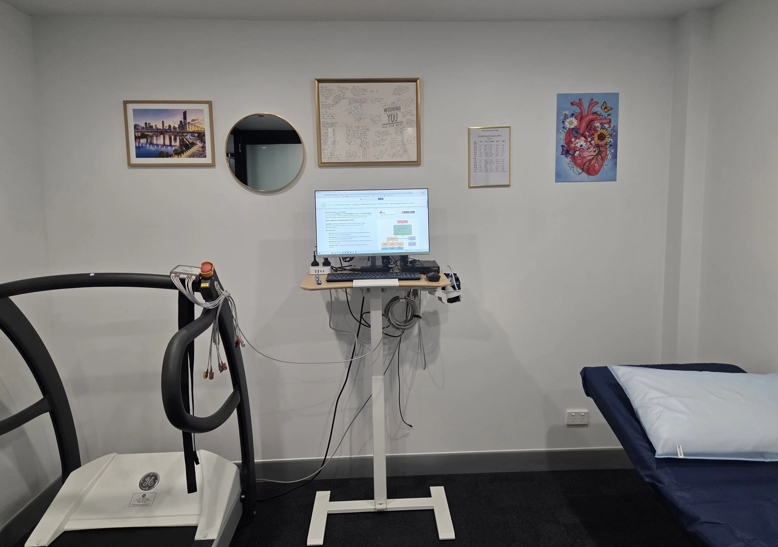 Hospital room with a treadmill, a computer monitor on a stand, a framed picture of a city skyline, a framed handwritten note, a heart anatomy poster, and a bed with a blanket.
