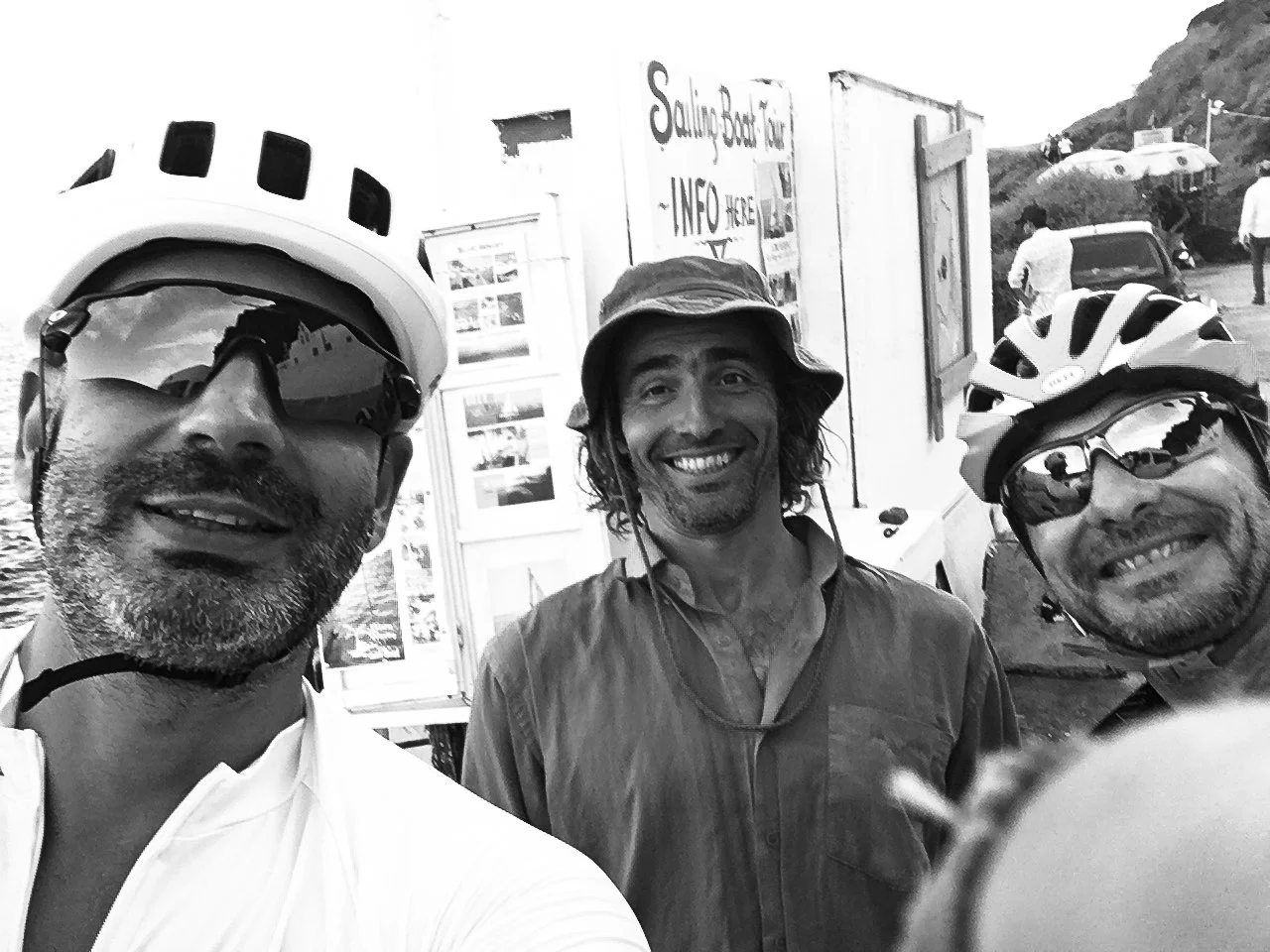 Three men wearing cycling helmets and sunglasses taking a selfie outdoors, with a signboard and other people in the background.