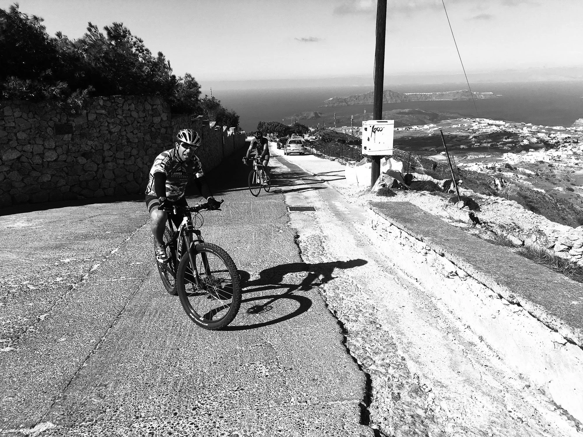 Two cyclists riding uphill on a winding road with a stone wall on the left and a steep drop on the right, overlooking a coastal landscape with water and land in the background, under a clear sky.