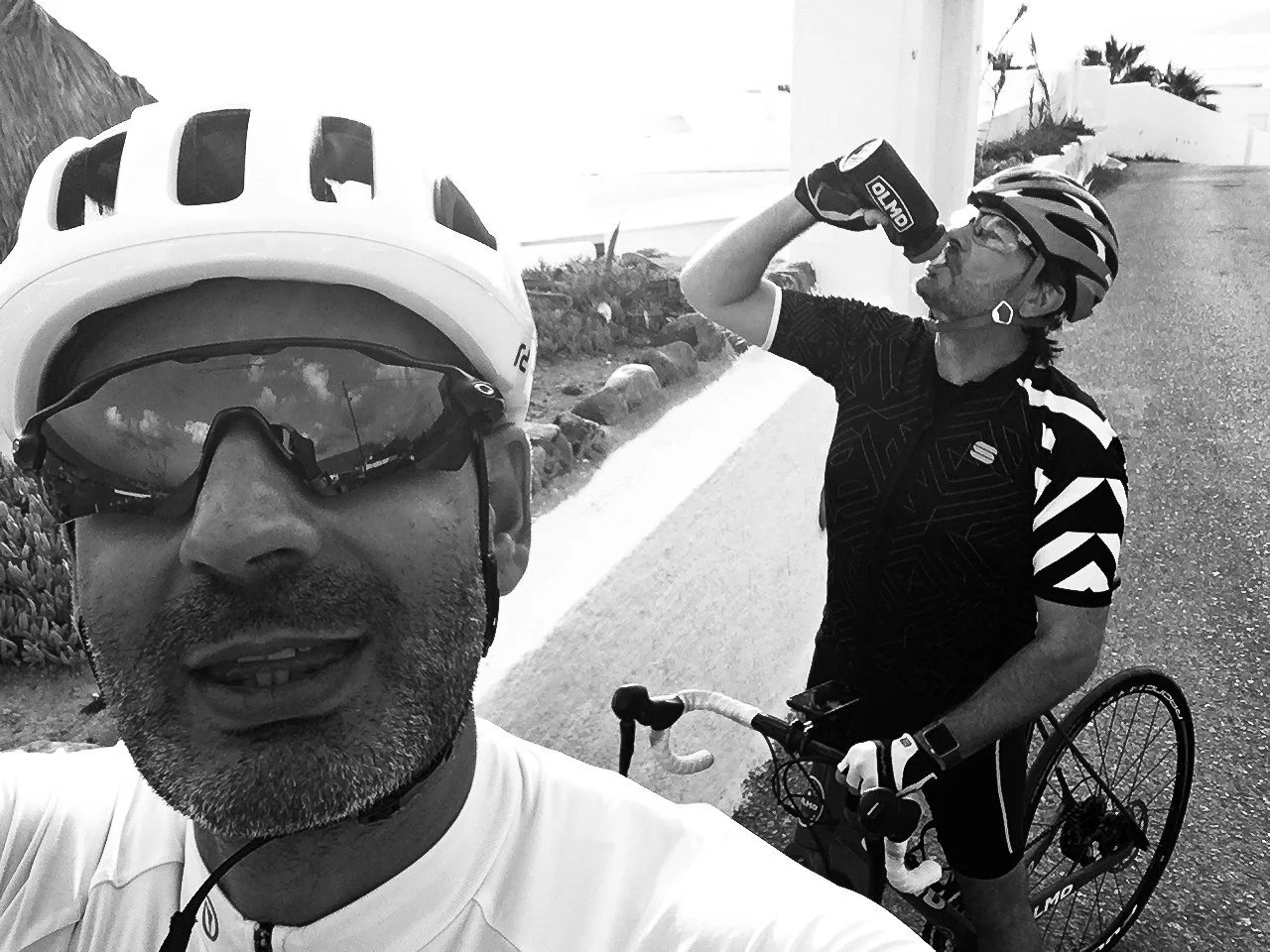 Two male cyclists wearing helmets and sunglasses, one drinking water and the other taking a selfie, outdoors on a paved road with plants and rocks in the background.