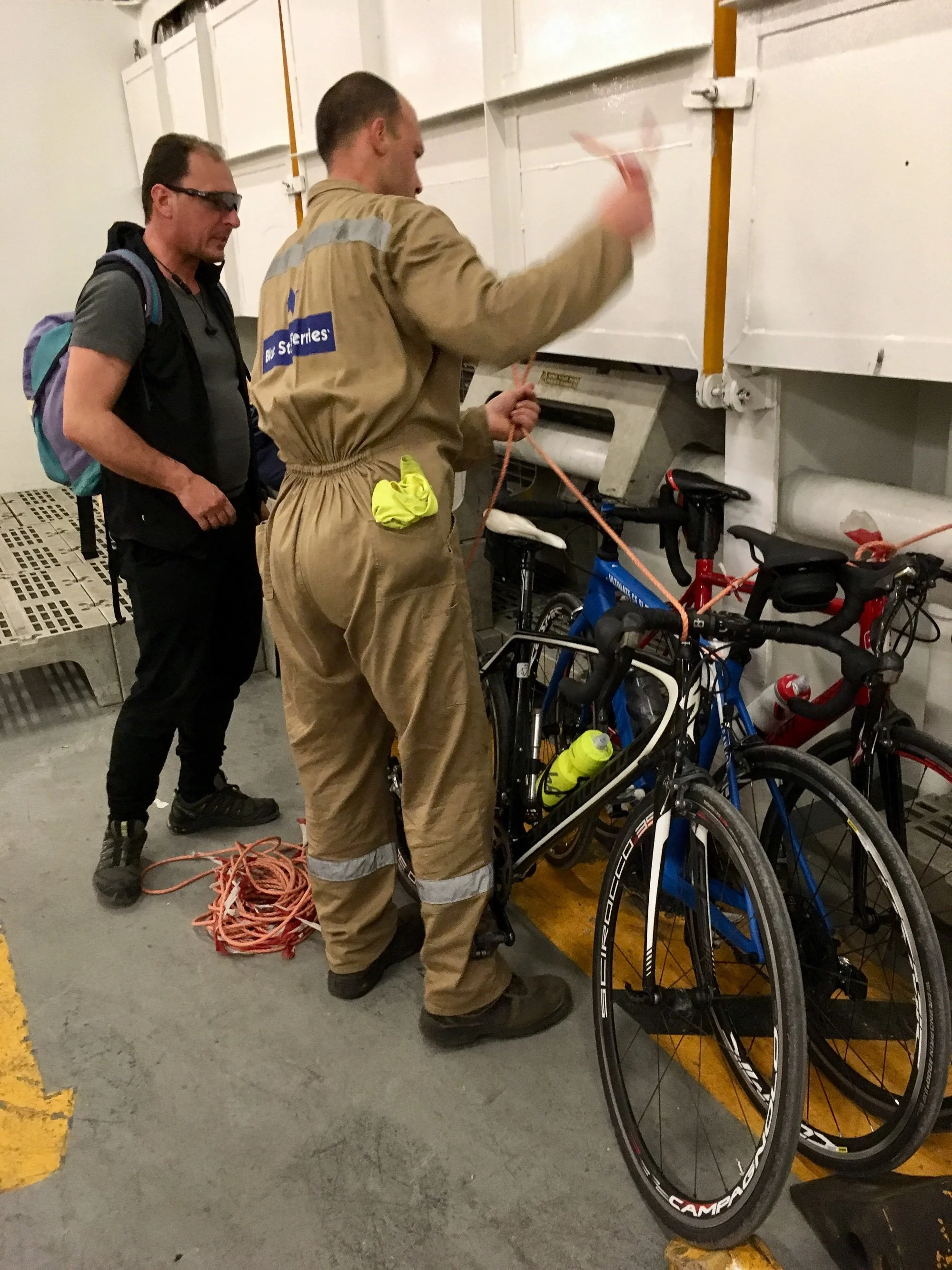 A firefighter in beige uniform is securing a bicycle with orange ropes, while another person with a backpack looks on inside a garage or storage area.