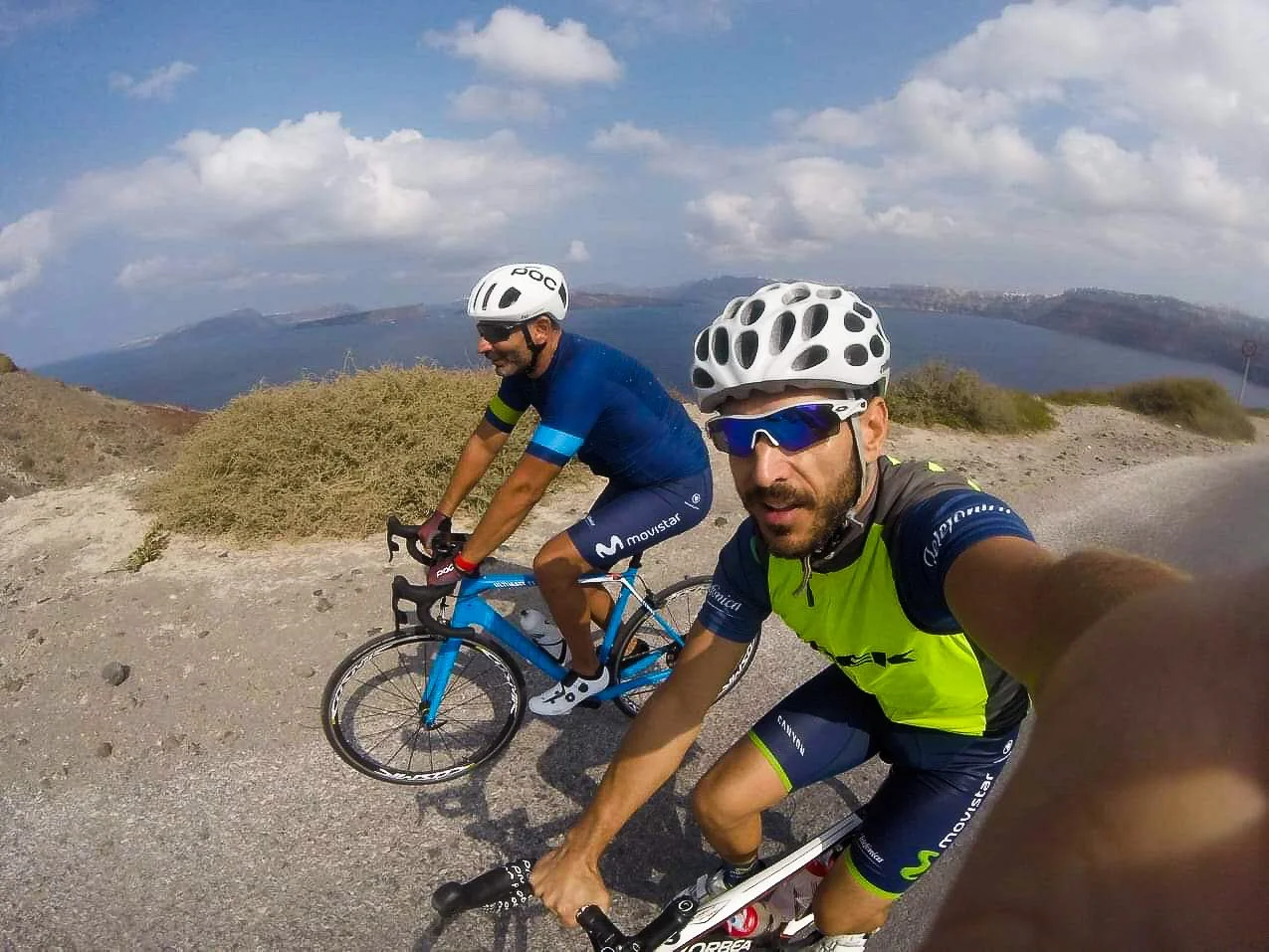 Two male cyclists riding bikes on a mountain road with a body of water and hills in the background, wearing helmets and cycling uniforms.