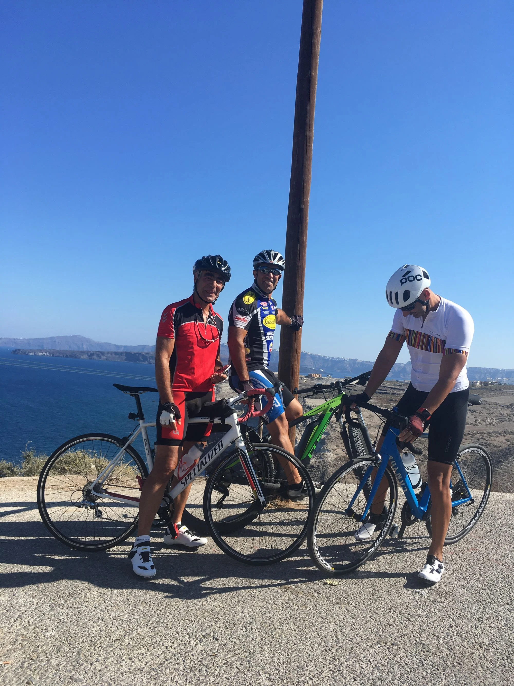 Three cyclists in helmets and athletic clothing with bikes near a body of water and rocky landscape under a clear blue sky.