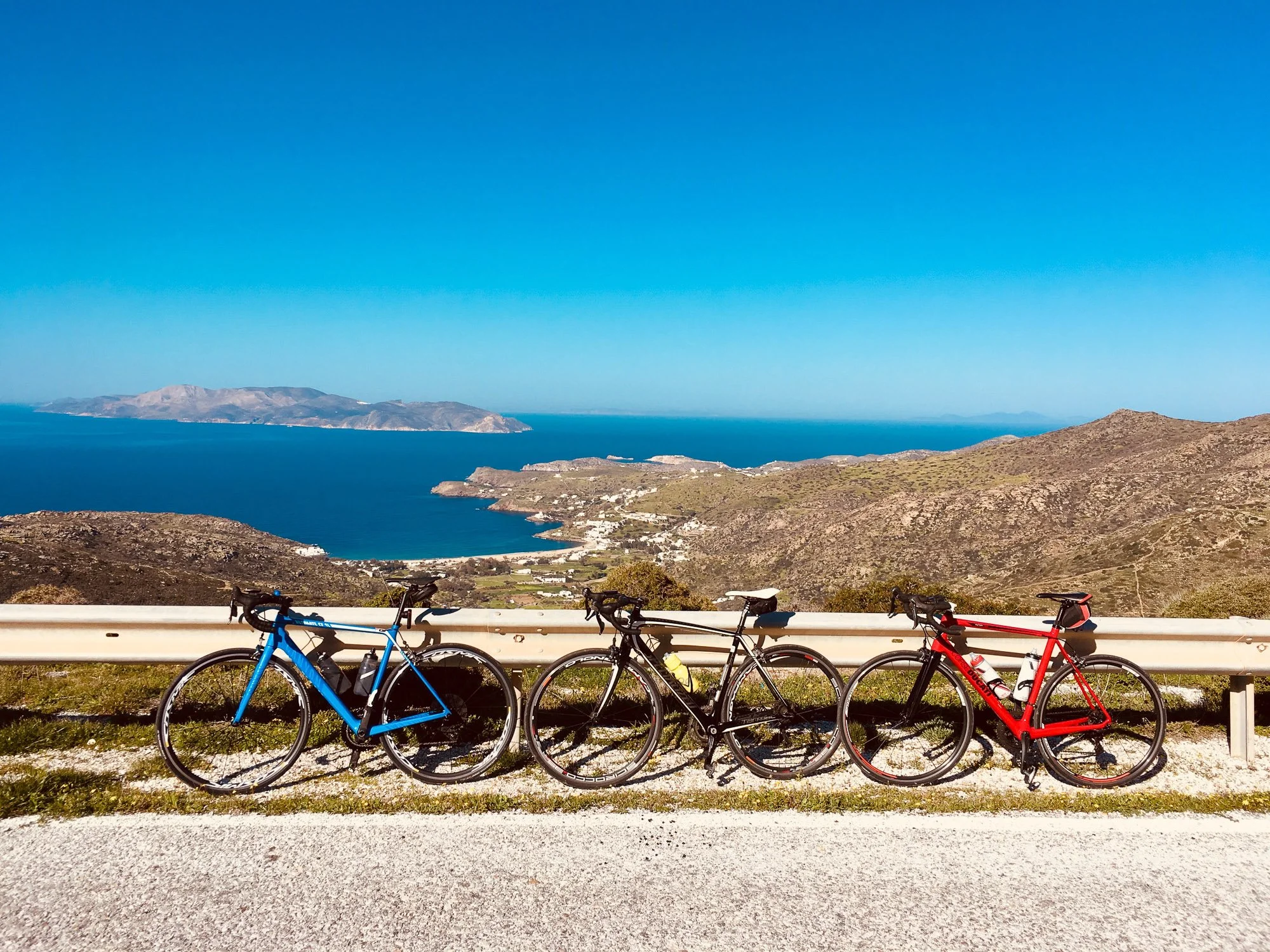 Three bicycles parked by a guardrail overlooking a scenic coastal landscape with hills, ocean, and distant islands under a clear blue sky.