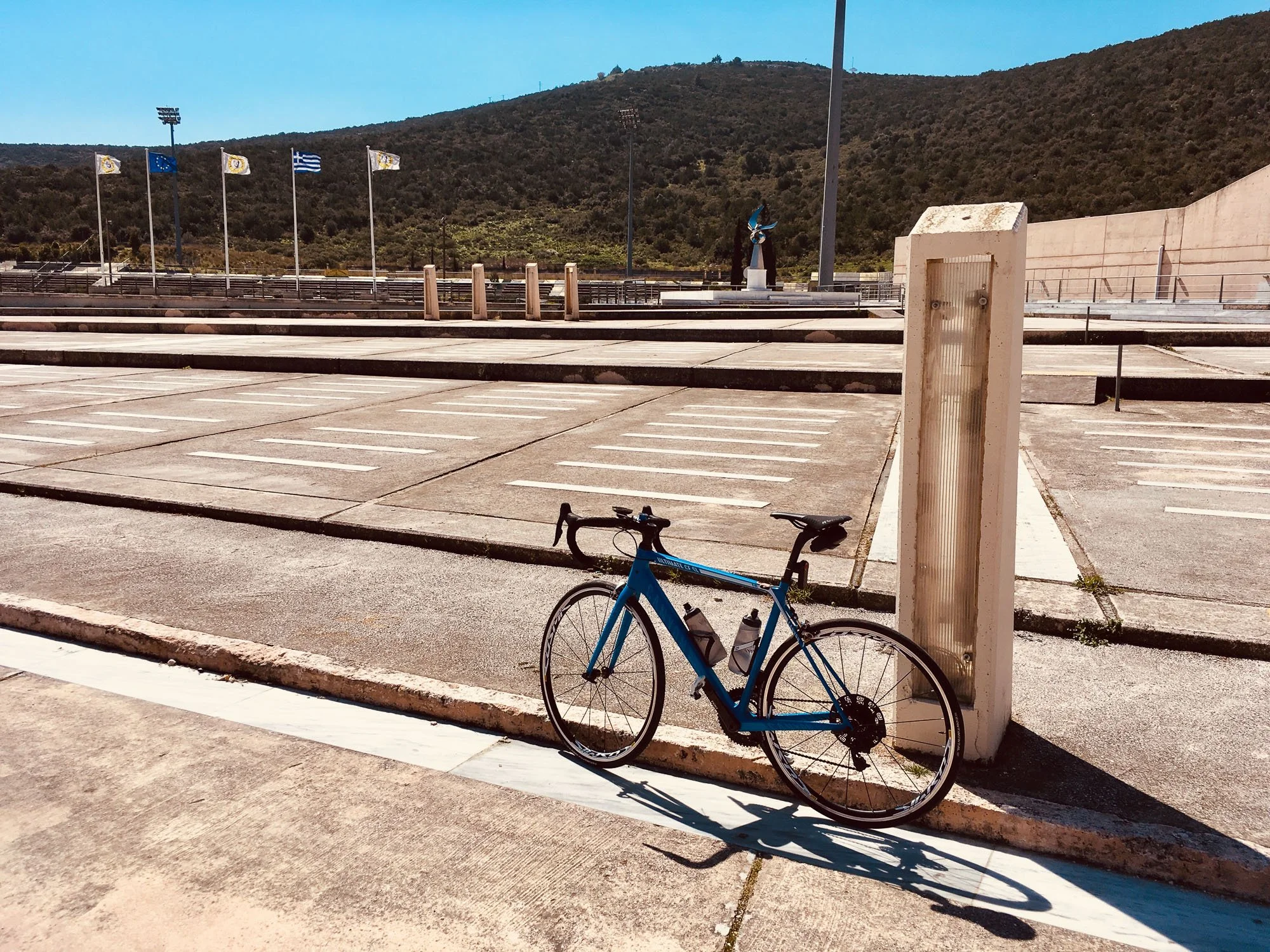 A blue road bike leaning against a barrier near an empty parking lot and a hillside in the background with flags flying.