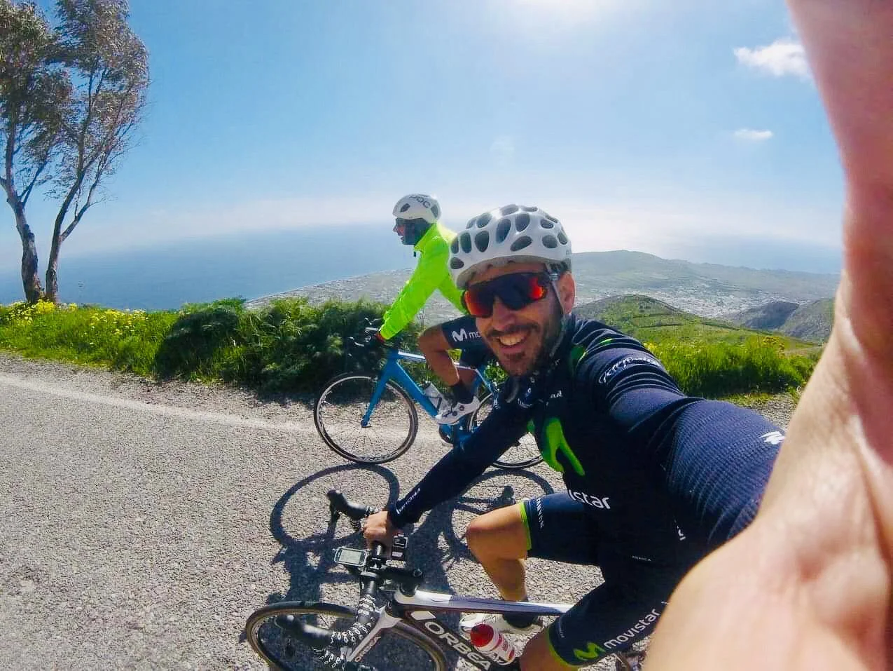Two cyclists wearing helmets and sunglasses riding on a mountain road with scenic ocean view in the background.
