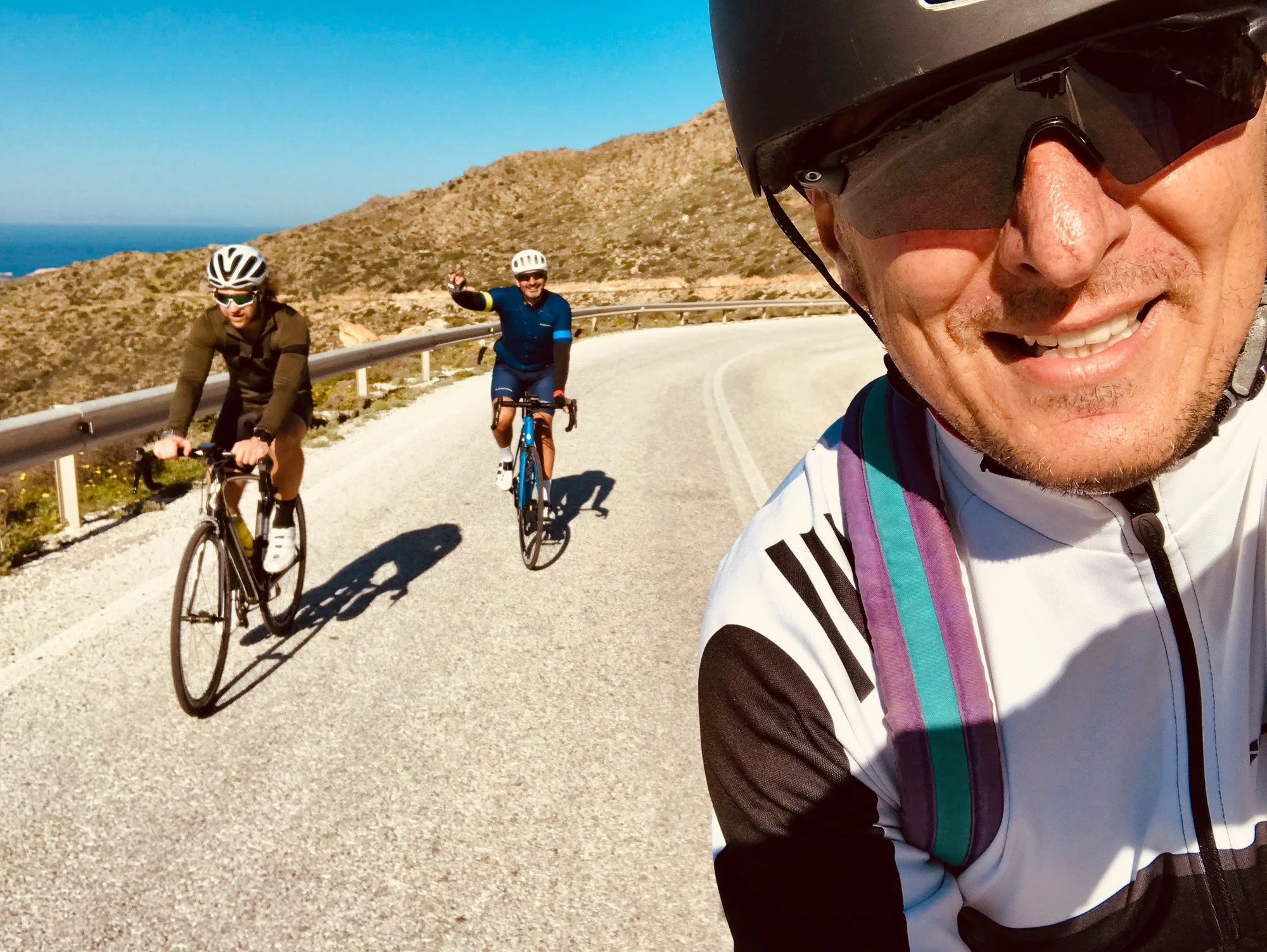 Three cyclists riding on a winding mountain road with a clear blue sky and rocky hills in the background, one of them taking a selfie.