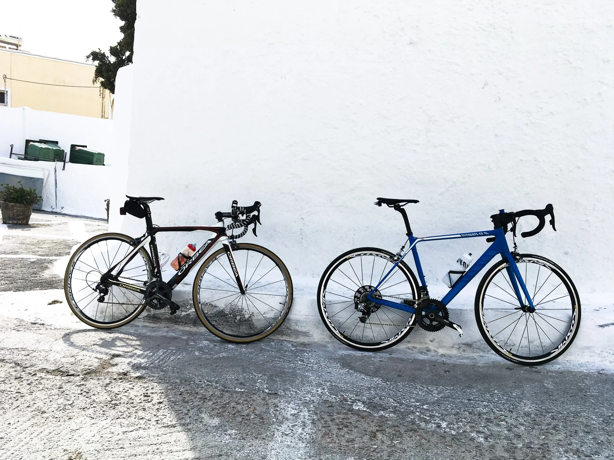 Two bicycles leaning against a white wall outside, with one bike being black and red and the other of blue color.