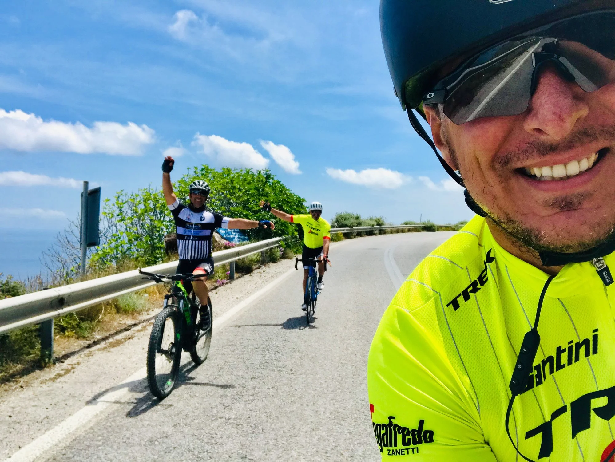 Man smiling with a helmet and yellow cycling jersey taking a selfie, two cyclists with helmets and jerseys riding on a mountain road with guardrail and greenery, clear blue sky with a few clouds in the background.