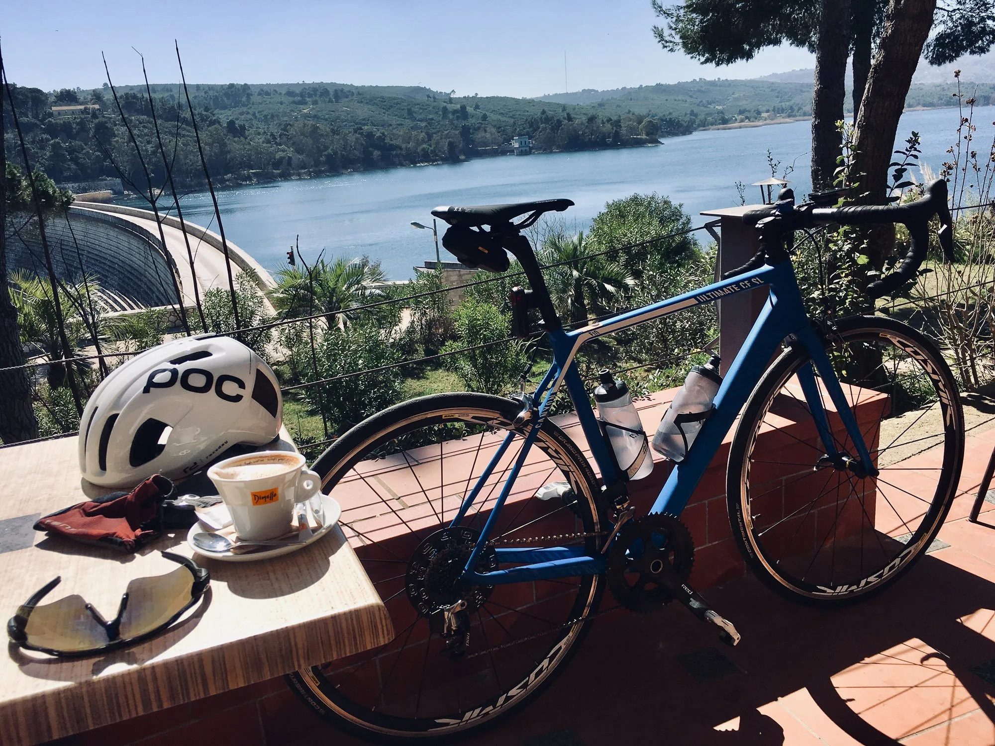 A blue road bicycle with water bottles, a white helmet, sunglasses, and a cup of coffee on a table overlooking a body of water and green hills.