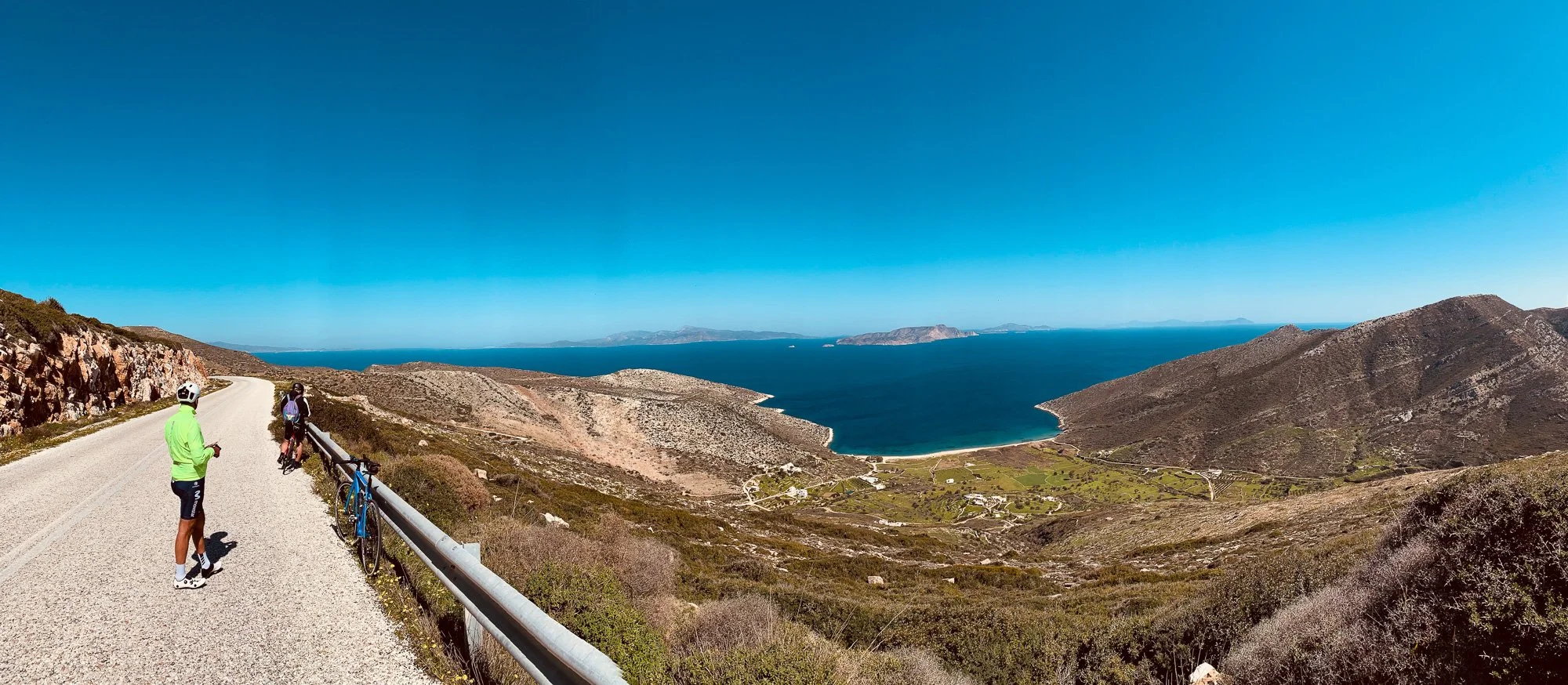Two cyclists and a pedestrian on a mountain road overlooking a large blue bay with small islands and hills under a clear blue sky.