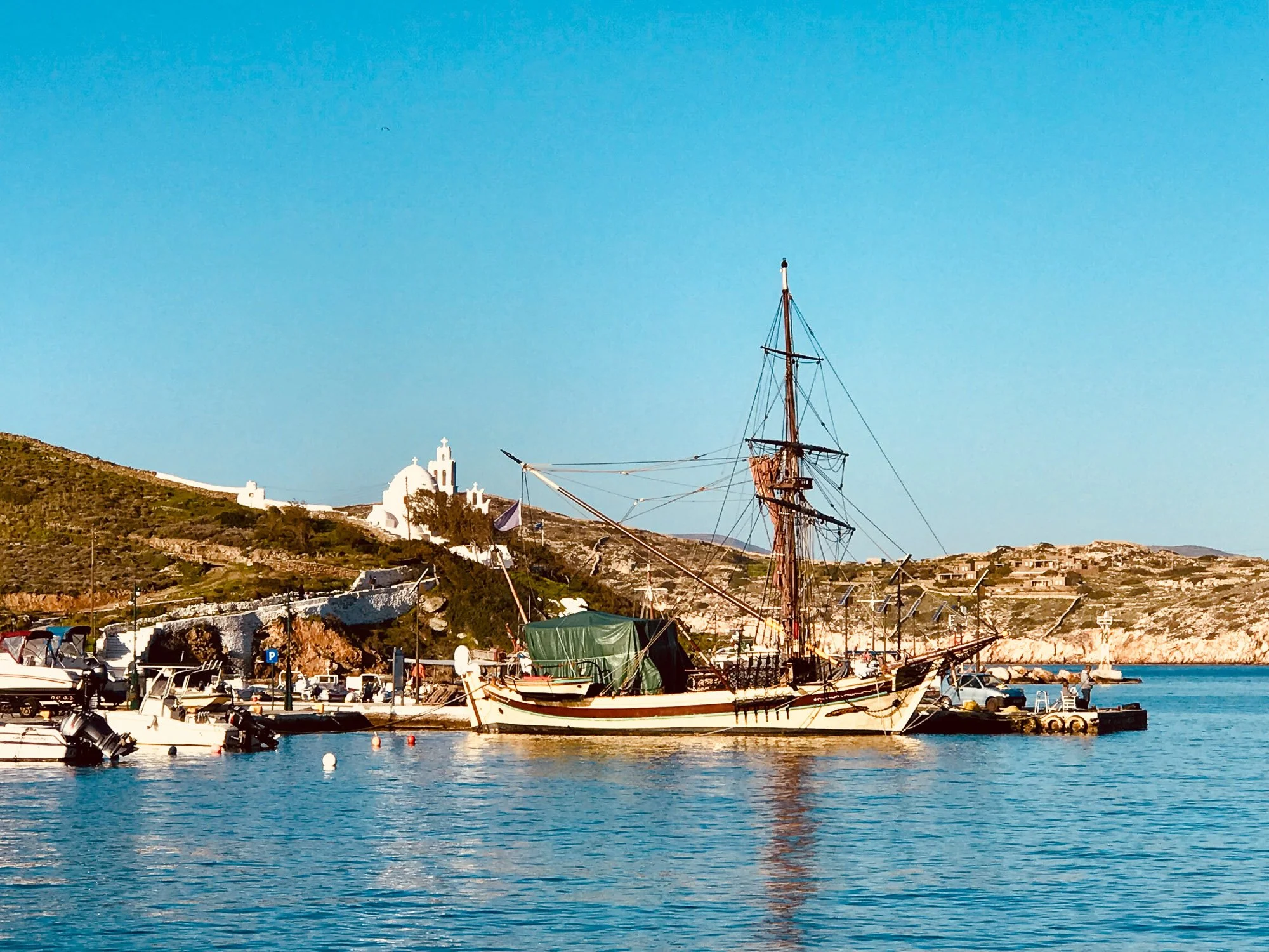A harbor with a large sailing ship anchored near the shore, smaller boats docked nearby, hillside with white buildings, and a blue sky.