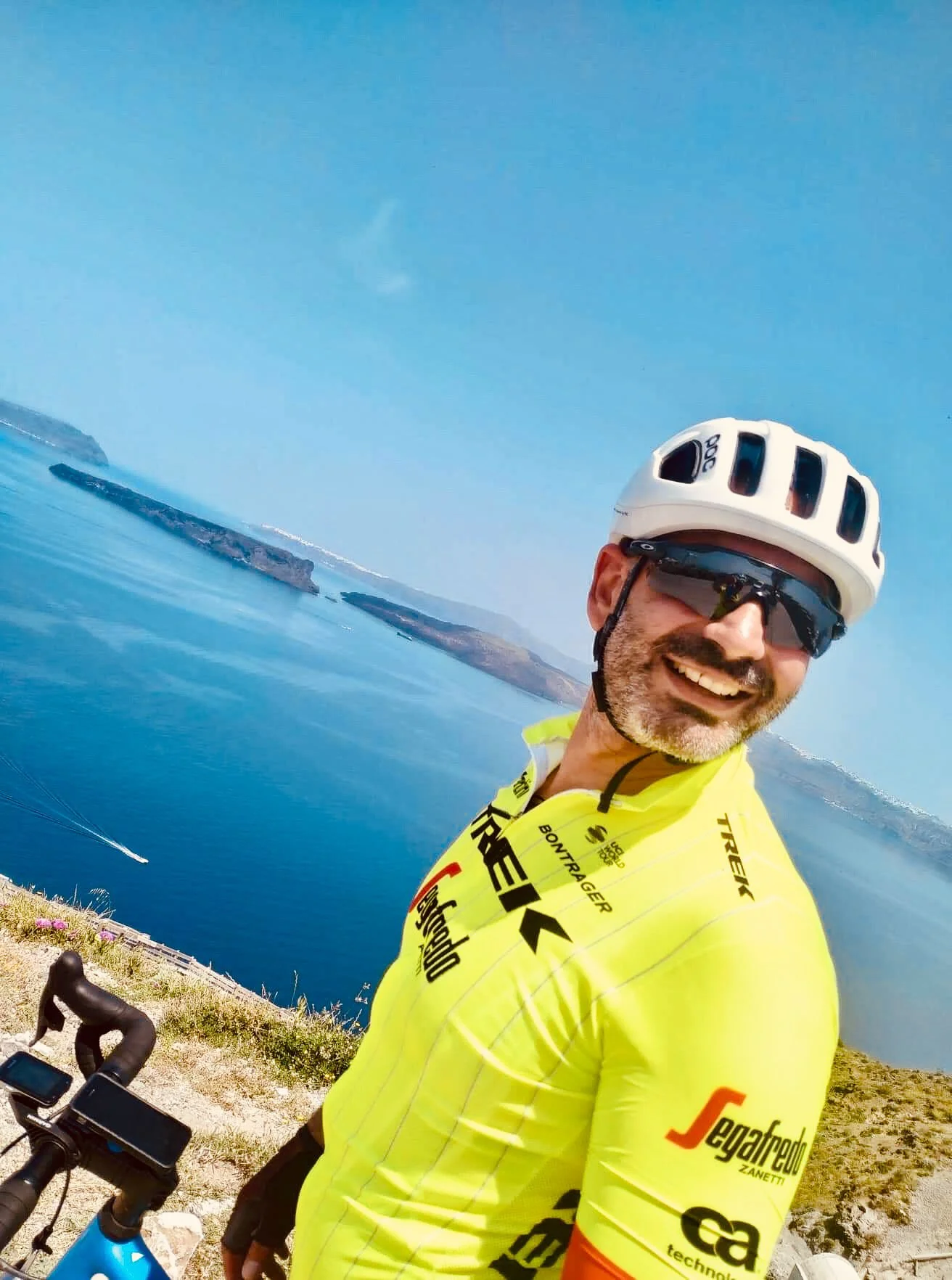 A man with a beard smiling, wearing a yellow cycling jersey, white helmet, and sunglasses, standing with a bicycle near a body of water with cliffs in the background.