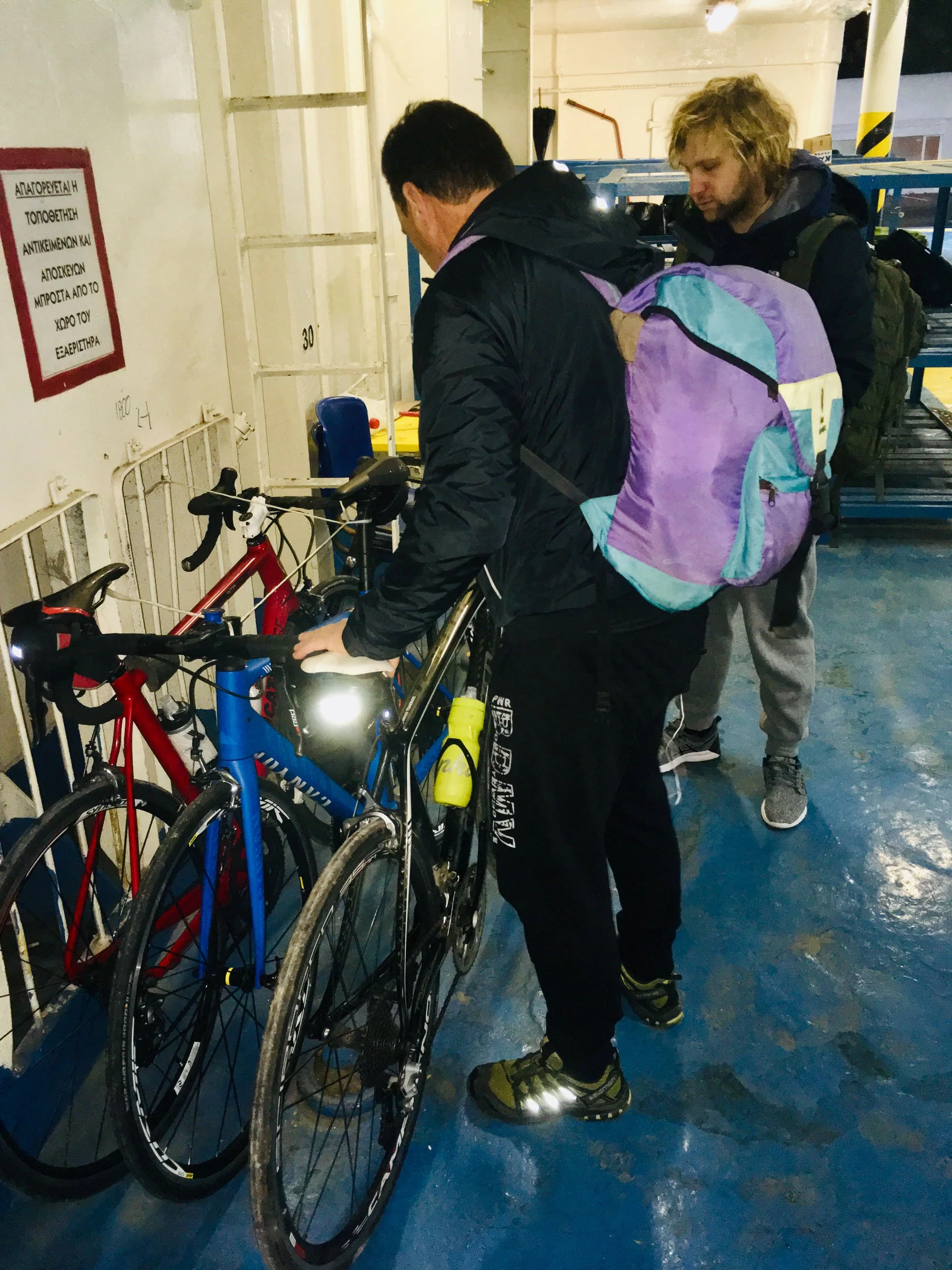 Two men standing with bicycles in an indoor parking area. One man is wearing a black jacket with a purple and turquoise backpack, and the other man has a gray hoodie and a dark jacket. The men are examining the bicycles.