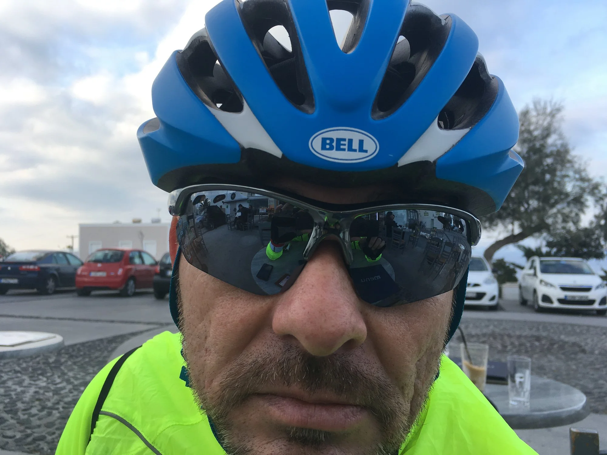Close-up of a man wearing a blue Bell bicycle helmet and reflective sunglasses, with a parking lot in the background and a drink on a table.