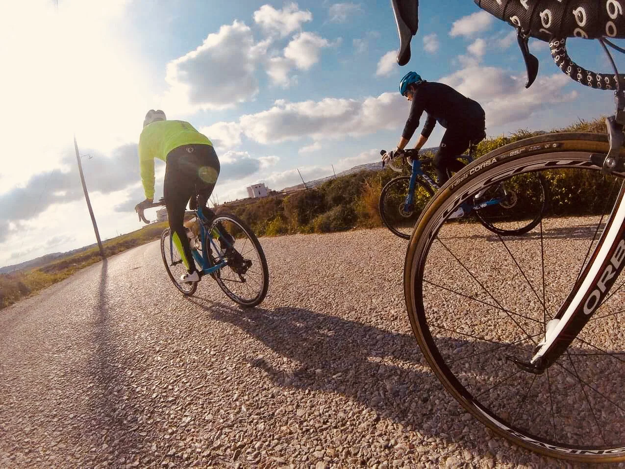 Two cyclists riding on a gravel road during daytime with a partly cloudy sky.