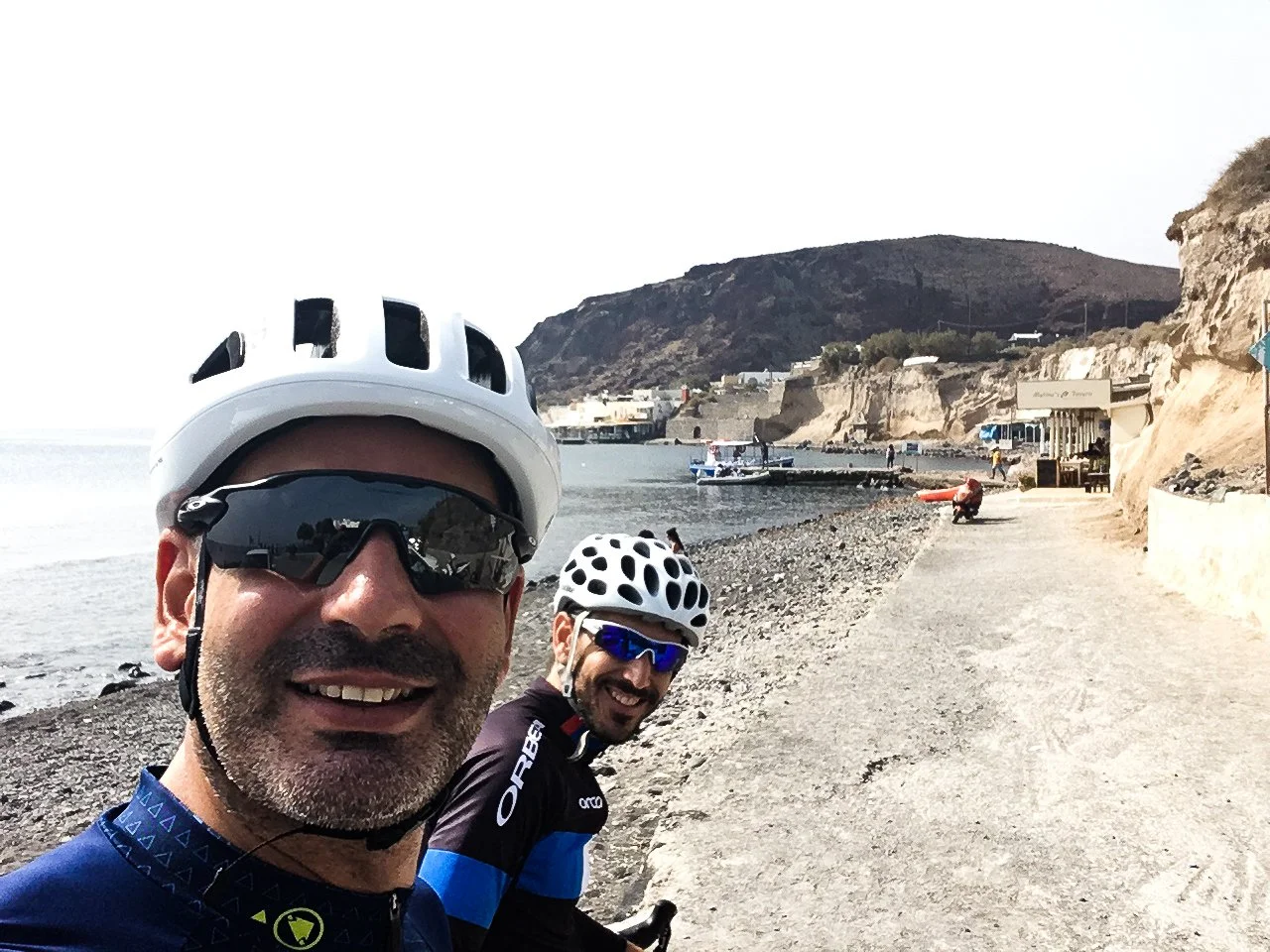 Two men in cycling gear and helmets taking a selfie on a coastal path with a pebble beach, boats, and cliffs in the background.
