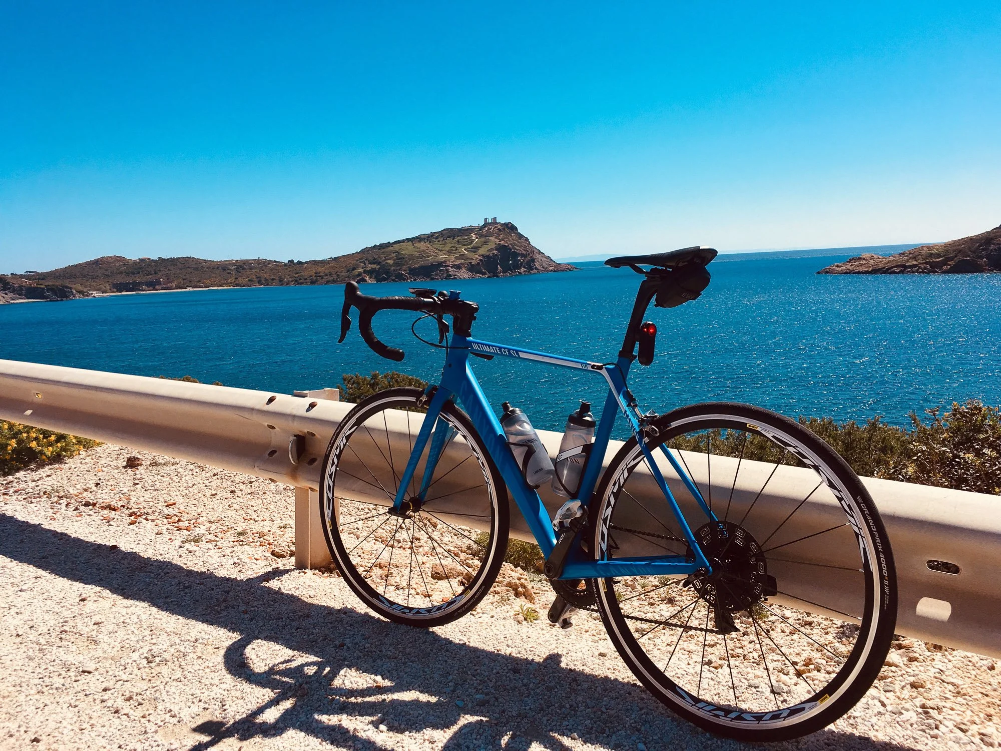 A blue road bike leaning against a white guardrail on a gravel road overlooking a large body of water with a small hill or island in the distance, under a clear blue sky.