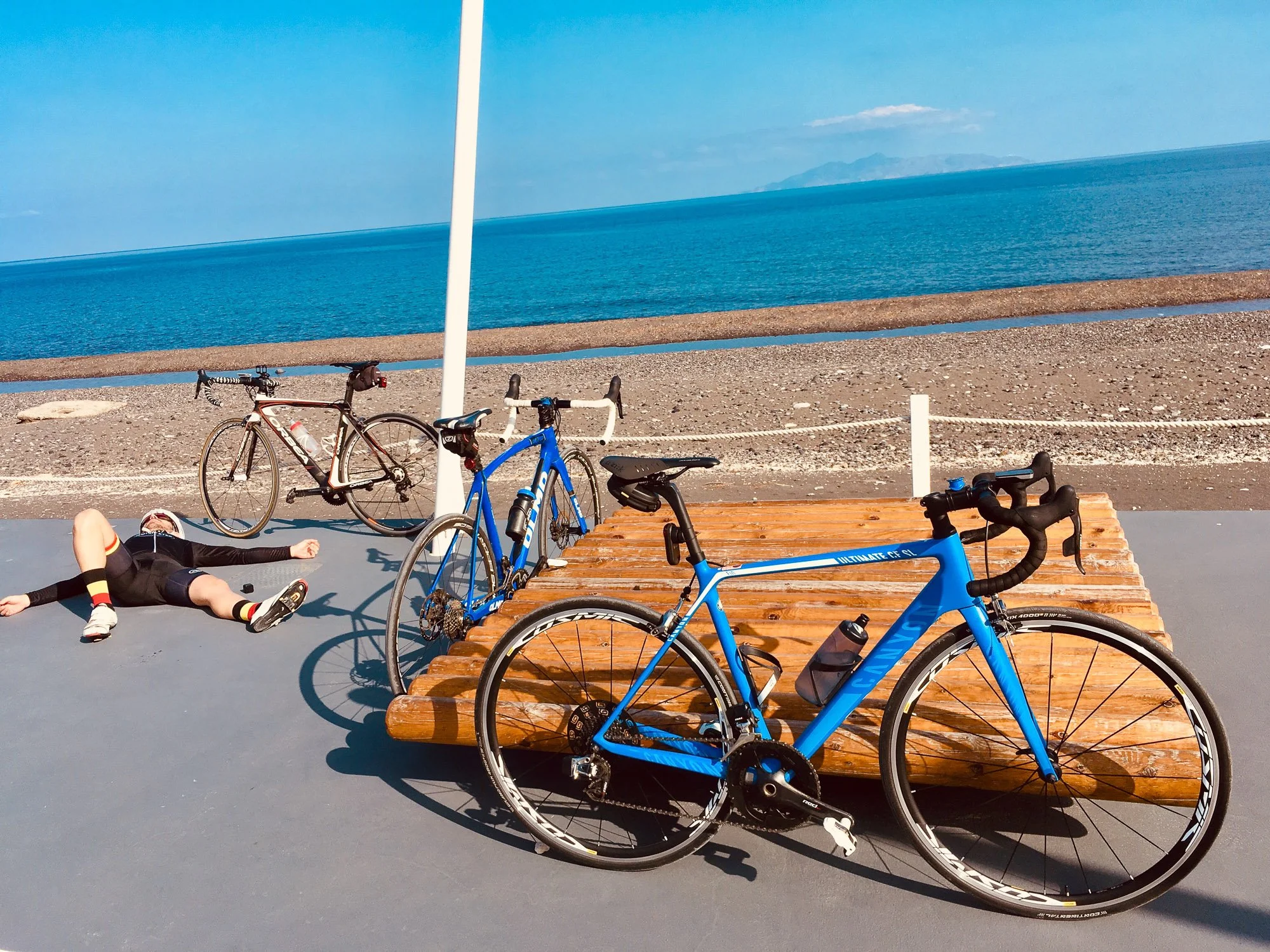 Two bicycles parked near a wooden bench at a seaside location, with a person lying on the ground, wearing cycling clothes, and an ocean view in the background.