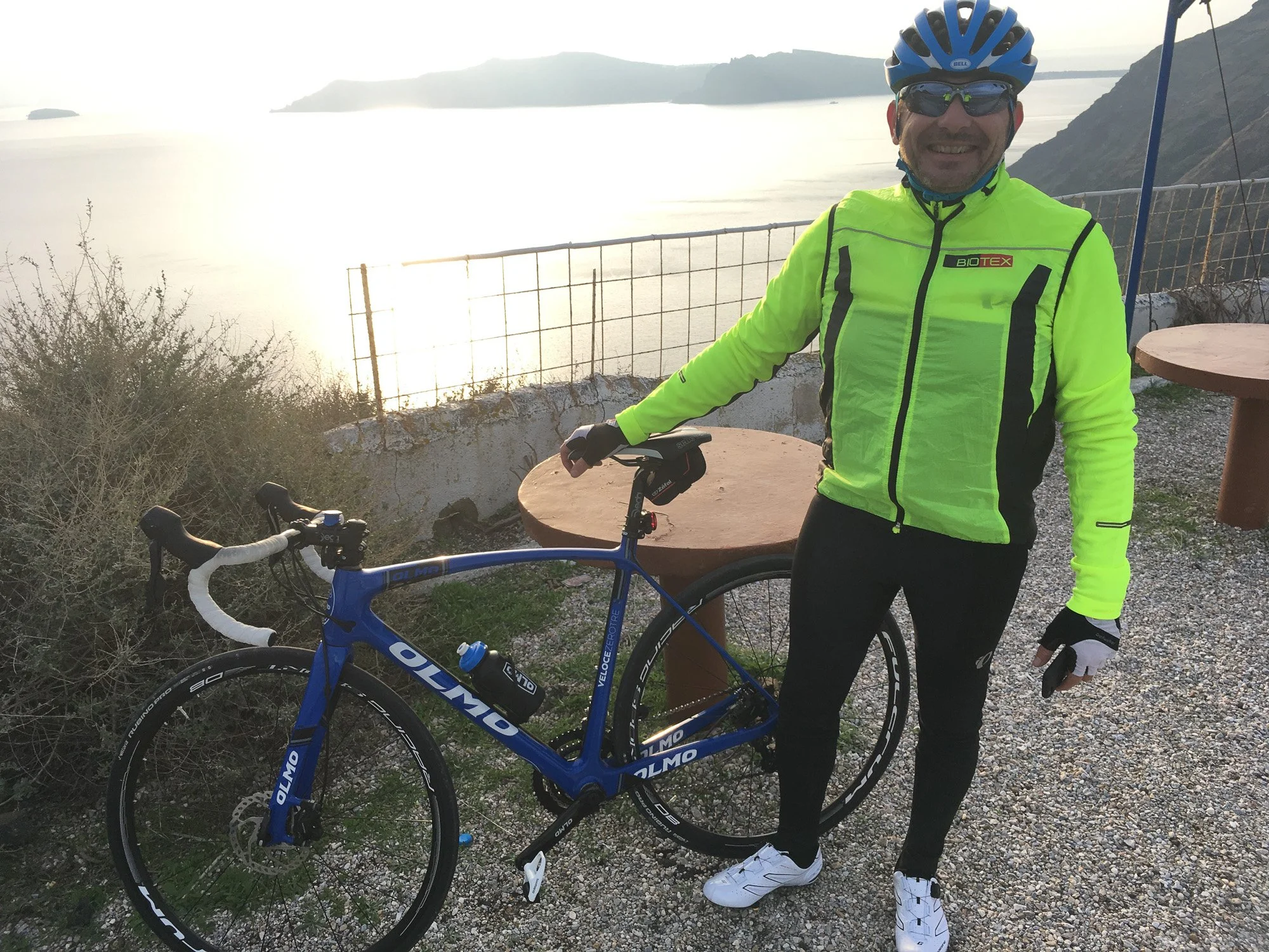 A man in cycling gear standing next to a blue road bike overlooking a body of water at sunset, with mountains in the background.