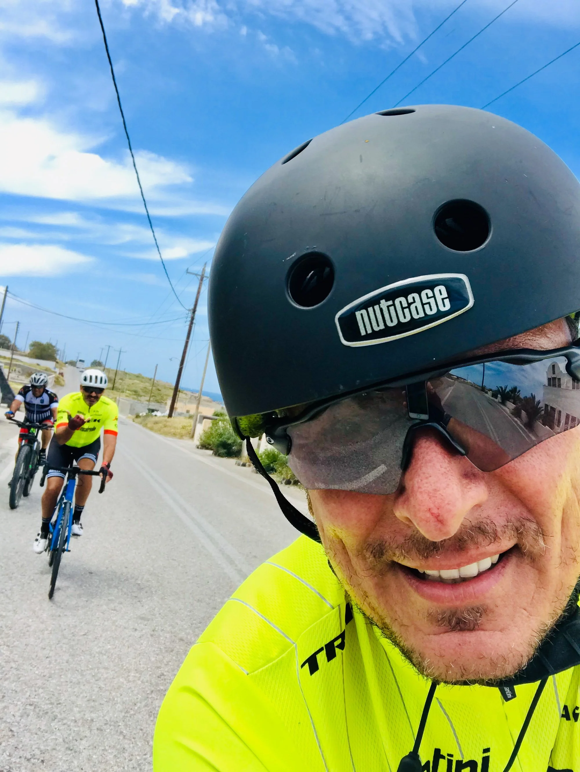 Close-up of a man wearing a black helmet with the logo 'nutcase' and dark sunglasses, smiling on a bicycle ride. In the background, three other cyclists are riding on a sunny road with blue sky and some clouds.