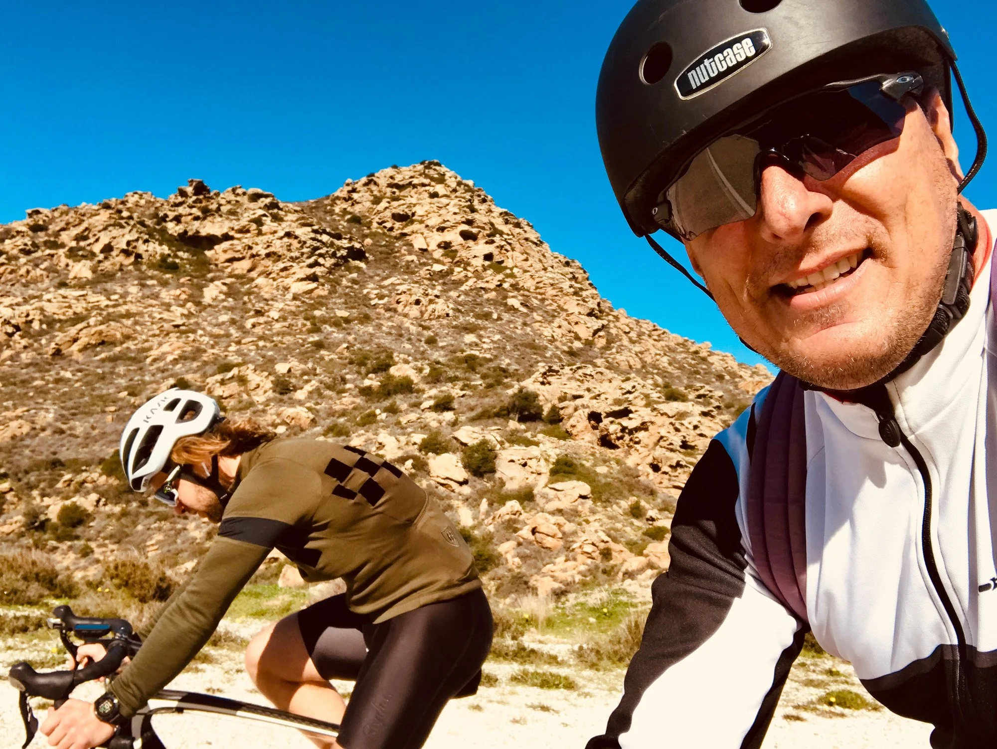 Two cyclists riding on a trail in a rocky desert landscape under a clear blue sky. The man in the foreground is taking a selfie, wearing a black helmet and sunglasses, smiling. The woman in the background is wearing a white helmet and a brown jacket,
