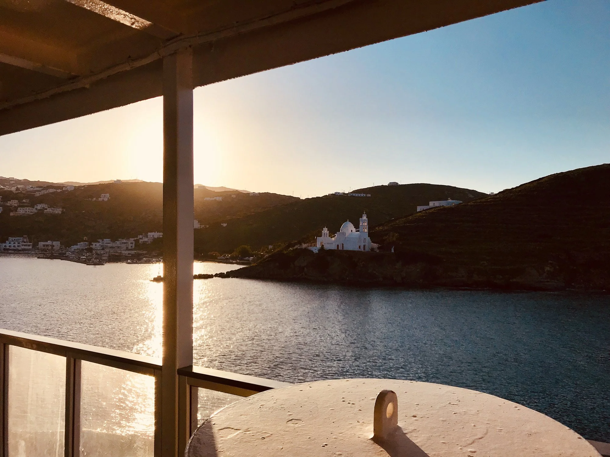 Sunset view of a lake with a white church on a hill and houses on surrounding hills, seen from a balcony with a table in the foreground.