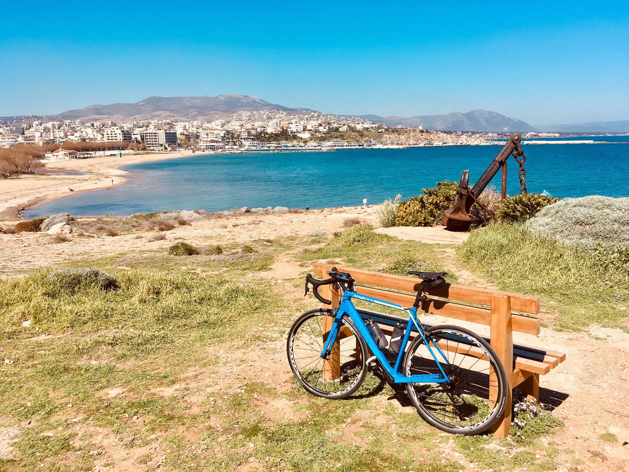 A scenic beach with a blue bicycle leaning on a wooden bench, overlooking the ocean with a cityscape and mountains in the background, and a large rusted anchor sculpture near the shoreline.