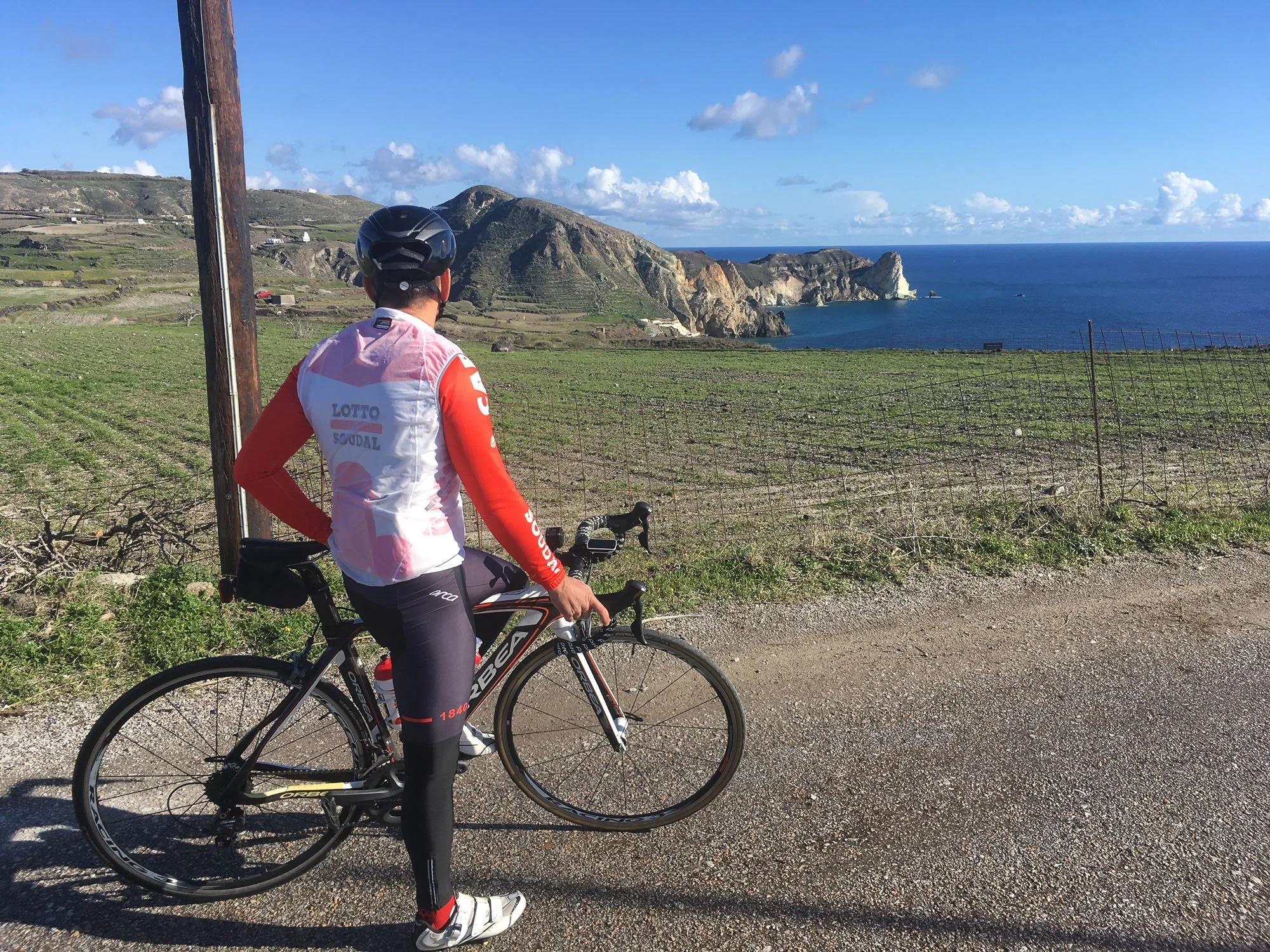A cyclist wearing a red and white cycling outfit and black helmet, standing next to a black road bike on a dirt path, with green fields, a fence, and cliffs overlooking the ocean in the background on a sunny day.